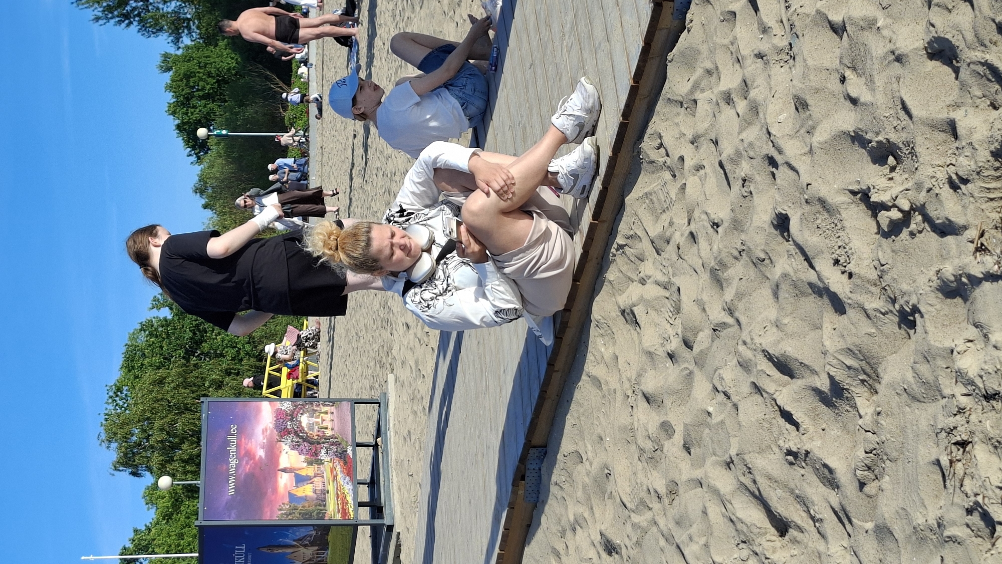 In Tallinn, Estonia, on a sandy beach with a wooden boardwalk, a woman is sitting on the boardwalk, looking at her phone. She has blonde hair tied in a bun, is wearing a white hooded jacket with a black graphic print, and light-colored shorts. She also has white headphones around her neck. To her right, a person wearing a blue baseball cap and a white t-shirt is sitting on the boardwalk. In the background, a large sign for "www.wagenkull.ee" is visible. Several other people are scattered across the beach and boardwalk, some sitting and others standing. Trees line the edge of the beach, and the sky is clear and blue.