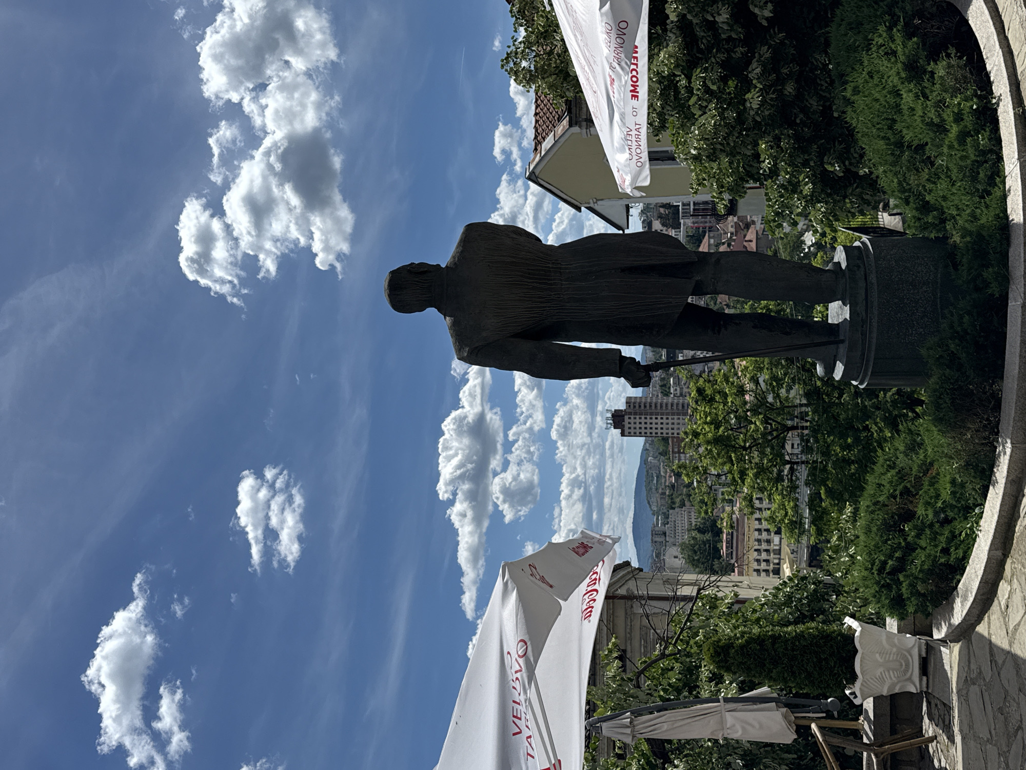 The image depicts a bronze statue of Stefan Stambolov in Veliko Tarnovo, Bulgaria. The statue is standing on a stone pedestal. The background consists of a partly cloudy blue sky and buildings of the city of Veliko Tarnovo. There are white umbrellas on either side of the statue. Trees are also visible.