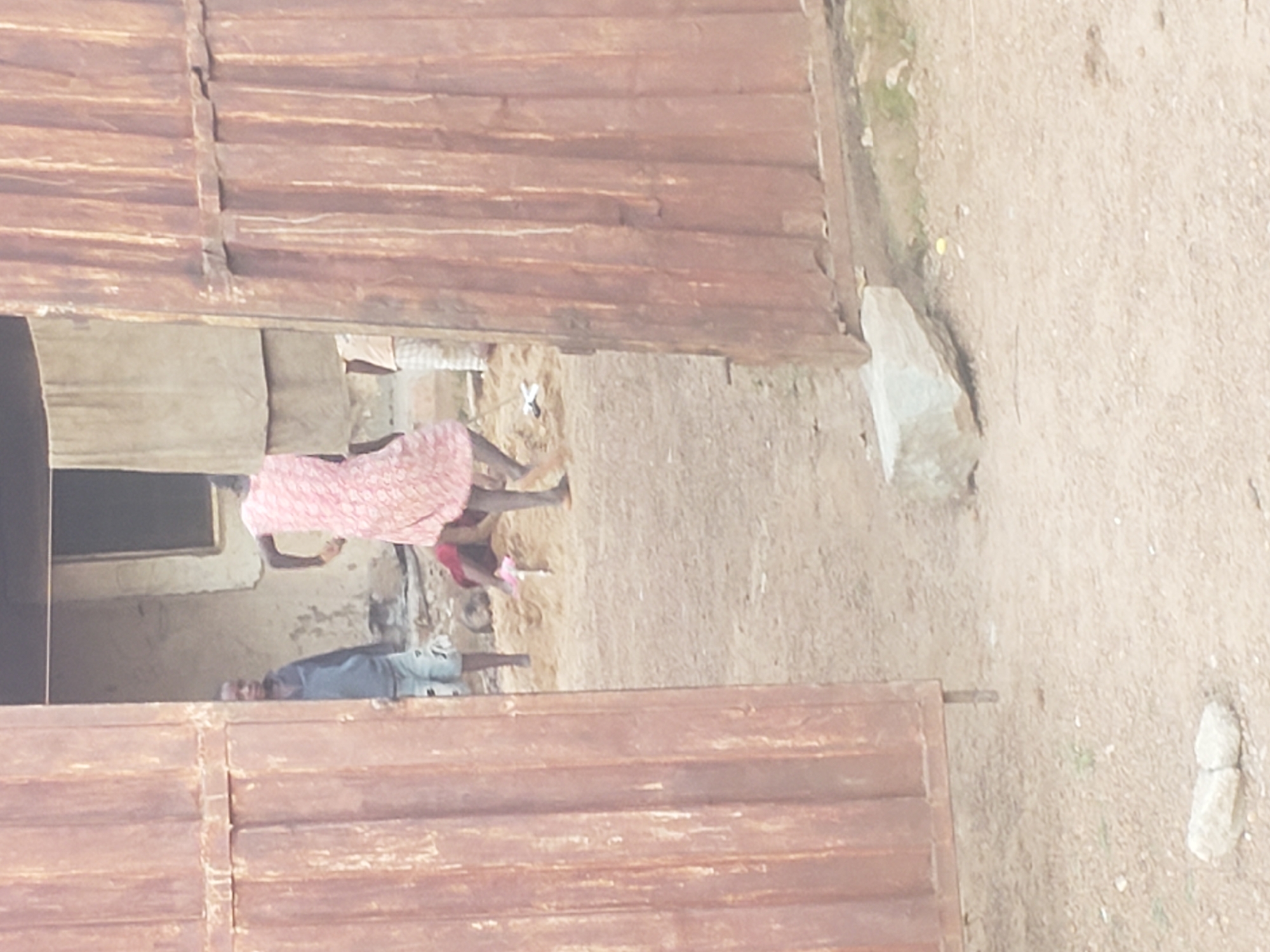 In Kachia, Nigeria, a group of children are playing in a sandy area in front of a building with corrugated metal doors. One child, wearing a pink dress, is bent over, appearing to be digging or playing with the sand. Another child, in a blue sweatshirt and shorts, stands to the left, observing. A third child, partially visible in the background, is also engaged in activity in the sand. The environment is dusty and appears to be an outdoor area with some vegetation. The building's exterior is weathered, suggesting it might be an older structure.
