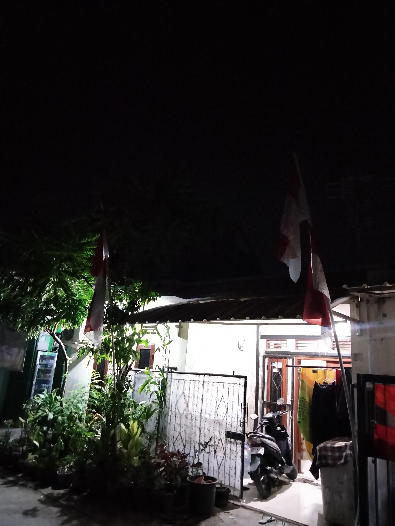 The image depicts a dimly lit scene in front of a modest dwelling, possibly in the city of Cariu, Indonesia. Two Indonesian flags are visible, one on the left and another on the right, hanging from poles. The flag on the left is partially obscured by lush green foliage, suggesting a garden or yard. The flag on the right is more prominent, showing its red and white bands.
In the foreground, a variety of potted plants are arranged, adding a touch of greenery to the scene. Behind the plants, a motorcycle is parked. The dwelling itself appears to have an open entrance or carport, with laundry hanging to dry. The overall atmosphere is one of a residential area at night, with ambient lighting contributing to the shadows and highlights. There are no people visible in the image.