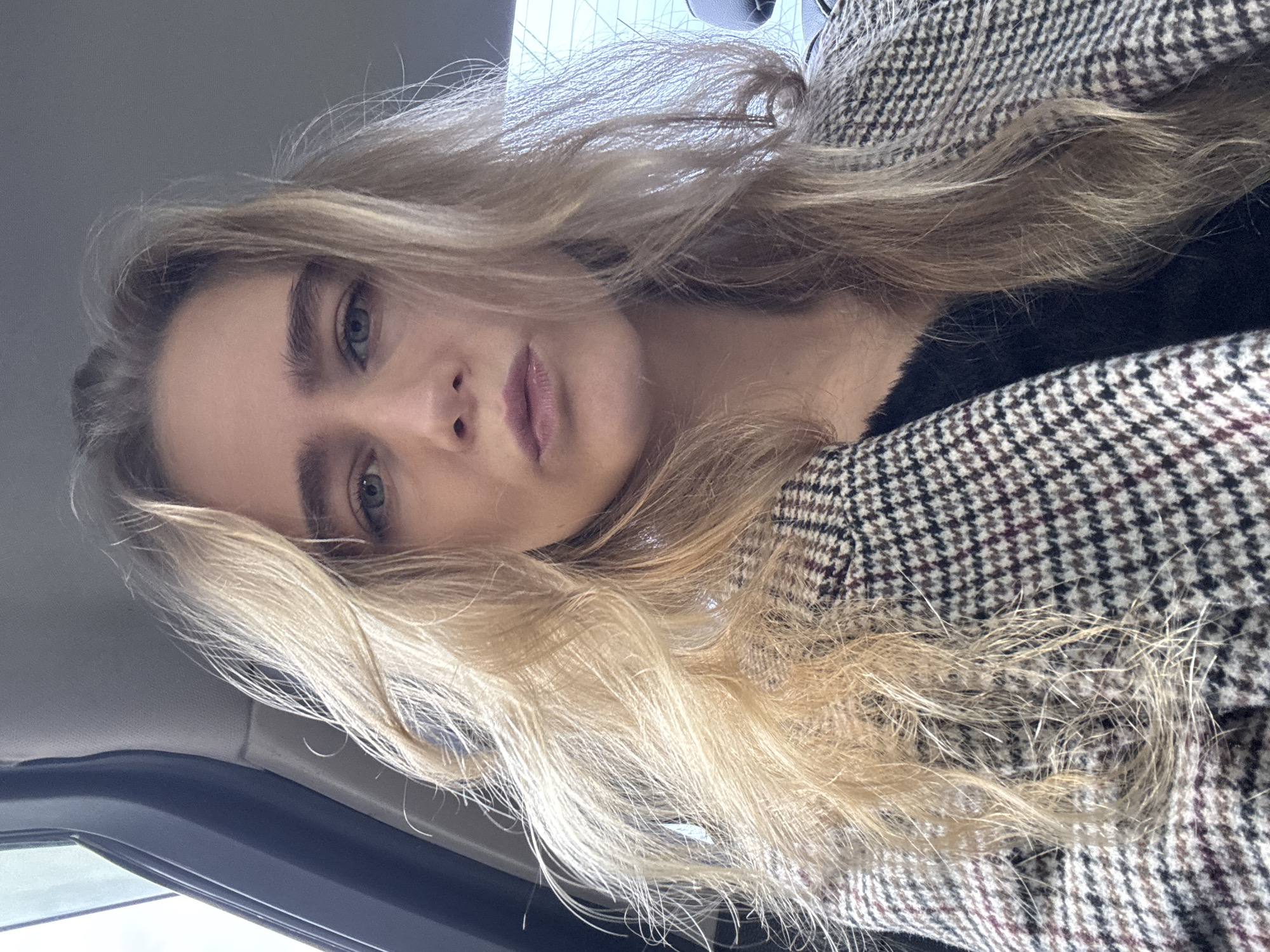 A close-up, angled shot shows a fair-skinned woman with blonde hair, wearing a tweed jacket, likely in a car. The location is unspecified.