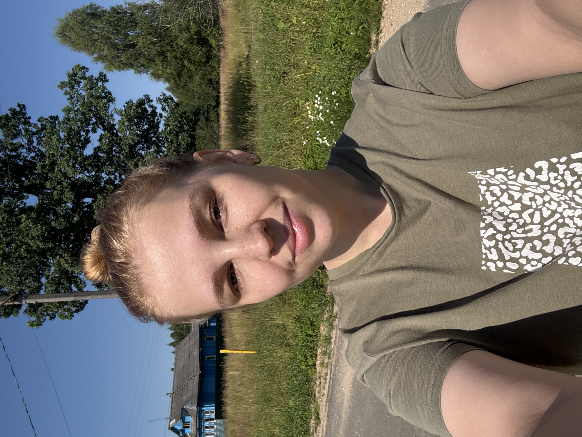 A young Caucasian woman with blonde hair pulled up in a bun is taking a selfie outdoors on a sunny day. She is wearing a green t-shirt with a white leopard-print design on the chest. The background shows a blue house with white trim, a tall green tree, and a field of grass. The scene appears to be in the village of Dunilovo, Russia.
