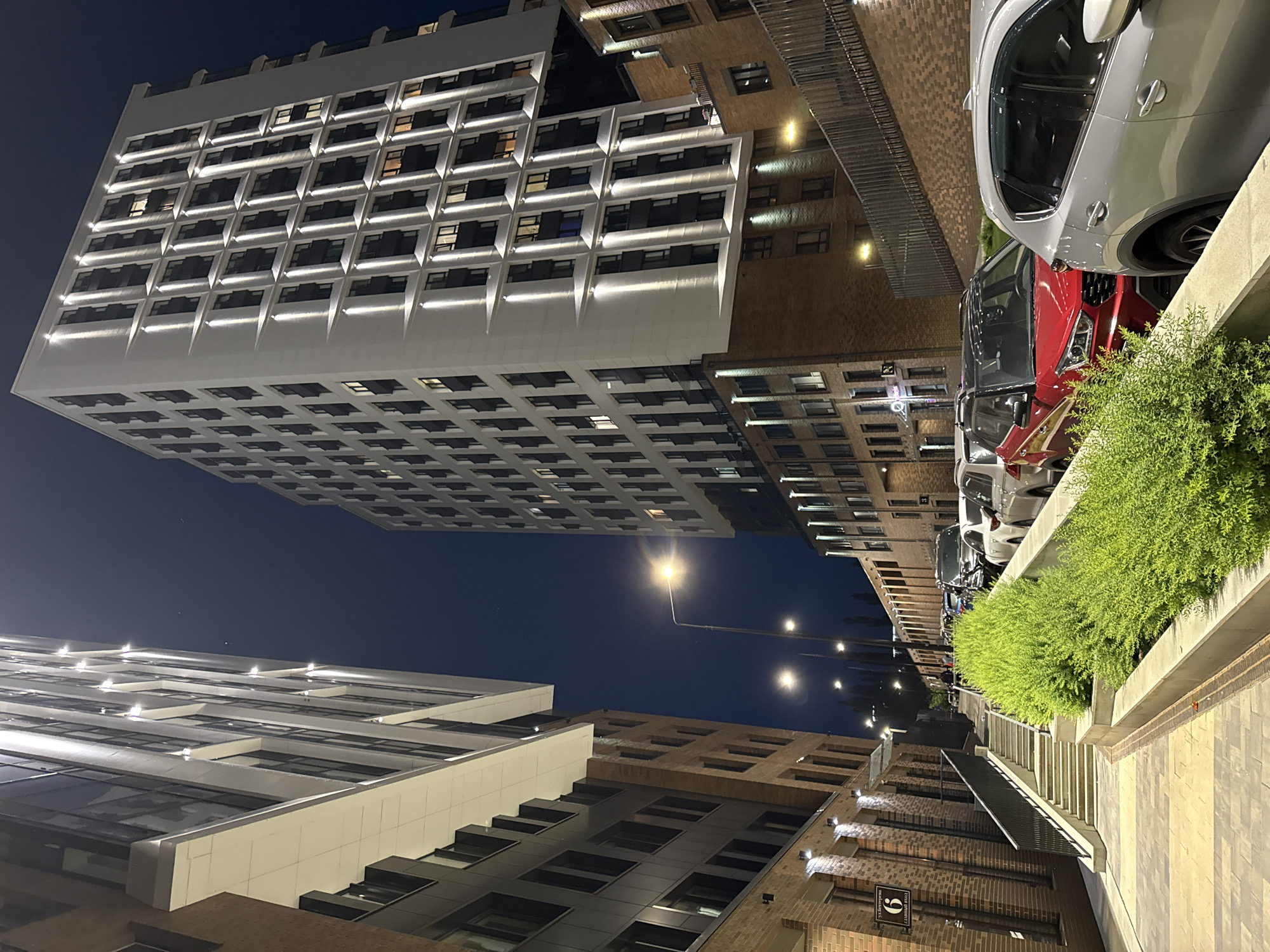 This nighttime image captures a modern urban street scene on Dezhurova Street in Ufa, Russia. The view is low-angle, looking up a paved pathway towards a street lined with multi-story buildings, parked vehicles, and illuminated landscaping.
To the left, a multi-story building features a brick facade on its lower levels, transitioning to dark gray panels and white horizontal bands on its upper floors. A white sign affixed to this building displays the number "6" and the Cyrillic text "УЛ. ДЕЖУРОВА" (Dezhurova Street) and "ГЛАВНЫЙ ВХОД" (Main Entrance). Adjacent to this building, a concrete staircase with a black metal handrail descends to the left.
The dominant structure is a tall, white multi-story building on the right, characterized by a modern facade. Its windows are arranged in vertical recessed sections, each individually illuminated by an uplight fixture that projects a defined beam onto the white building surface. Some windows display interior lighting. The base of this building also incorporates brickwork with smaller, wall-mounted lights.
In the foreground, a paved walkway, composed of yellow and gray blocks, leads into the scene. Along the right edge of this walkway, a raised concrete planter contains lush, vibrant green shrubbery. Beyond the planter, several vehicles are parked parallel to the curb, including a red SUV with a black roof and a silver SUV. Multiple streetlights, emitting bright yellow-white light, illuminate the street as it recedes into the background. The sky is dark blue. No individuals are clearly visible.