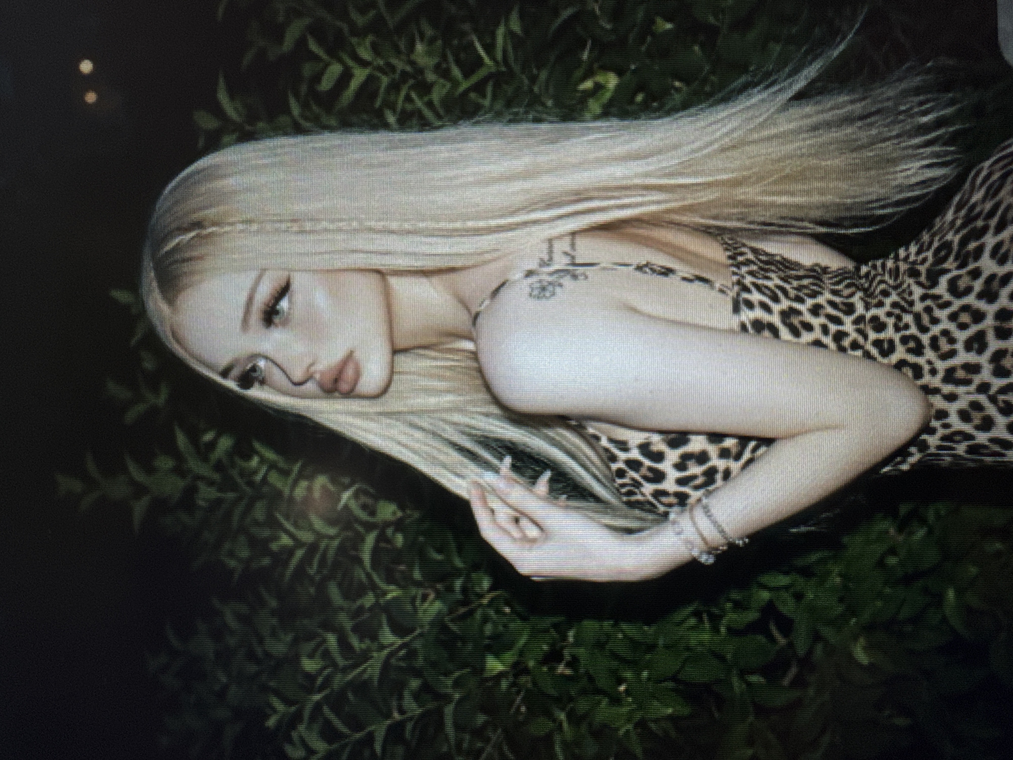 A young woman with very long, straight blonde hair, styled with thin braids near her forehead, is depicted in what appears to be an evening or nighttime setting. She is wearing a leopard print top or dress. Her head is tilted, and her face is turned towards the left side of the frame, showing a pensive or melancholic expression with open eyes and slightly parted lips.
A small, dark, stylized tattoo or design is visible on her right shoulder/upper arm. She also wears a bracelet on her left wrist. Her hands are gently posed, one near her chest and the other resting towards her hip.
The scene is set against a dark background dominated by lush green foliage, suggesting an outdoor location like a garden or a heavily planted area. Two faint, circular out-of-focus light sources (bokeh) are present in the upper left corner, indicating distant artificial lighting. The overall ambiance is atmospheric and serene, with the dark background contrasting with the woman's light hair and skin. The image exhibits a slightly grainy or pixelated quality, suggesting it may be a photograph of a screen or processed with a specific aesthetic filter. There is no clearly legible text.