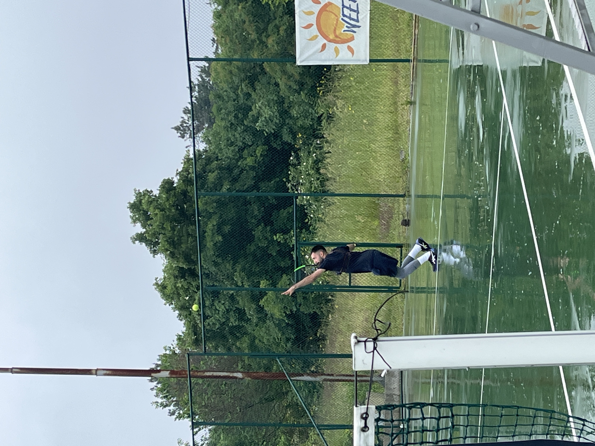 A man plays tennis outdoors, likely in Sozopol, Bulgaria. The scene is on a green tennis court, with lush green trees and a fence in the background. A banner with a sun emblem is affixed to the fence. The court appears wet, possibly from recent rain. The man is reaching with his arm extended as if in the middle of a swing. He is wearing black tennis clothing, with gray leggings and dark shoes. A tennis ball is positioned in the air near the fence. A tennis net and post is visible. The sky is overcast.