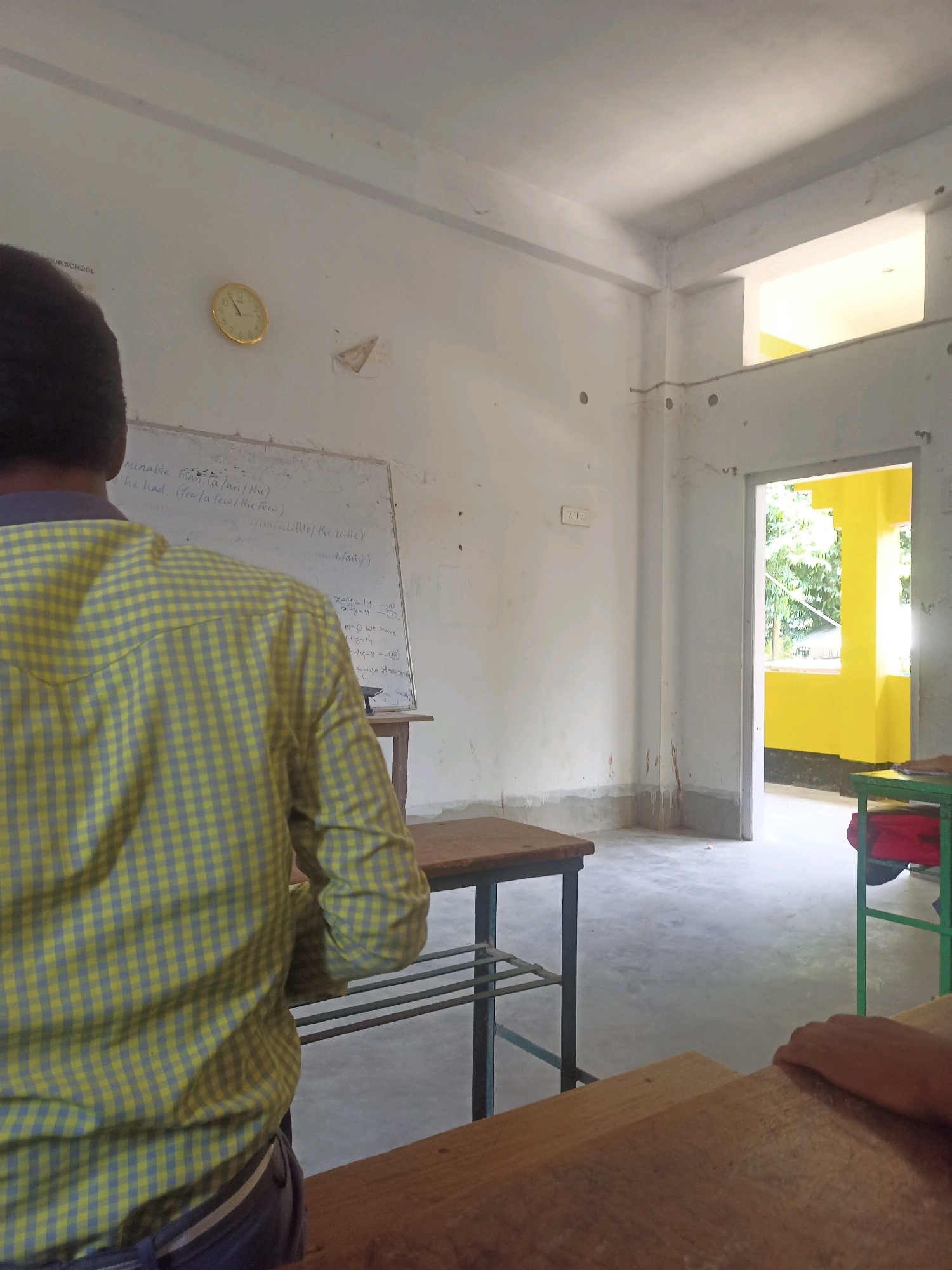 The image depicts the interior of a classroom in Hatsinigmari, India. A student, seen from behind, is wearing a yellow and grey checkered shirt and dark pants. They are facing a whiteboard with some writing and mathematical equations. In the background, a round analog clock is mounted on the wall. The classroom has simple wooden desks and a door opening to a brightly lit area with yellow accents. The walls are plain and show some signs of wear. The flooring appears to be concrete. Sunlight streams in from the doorway, illuminating the space.