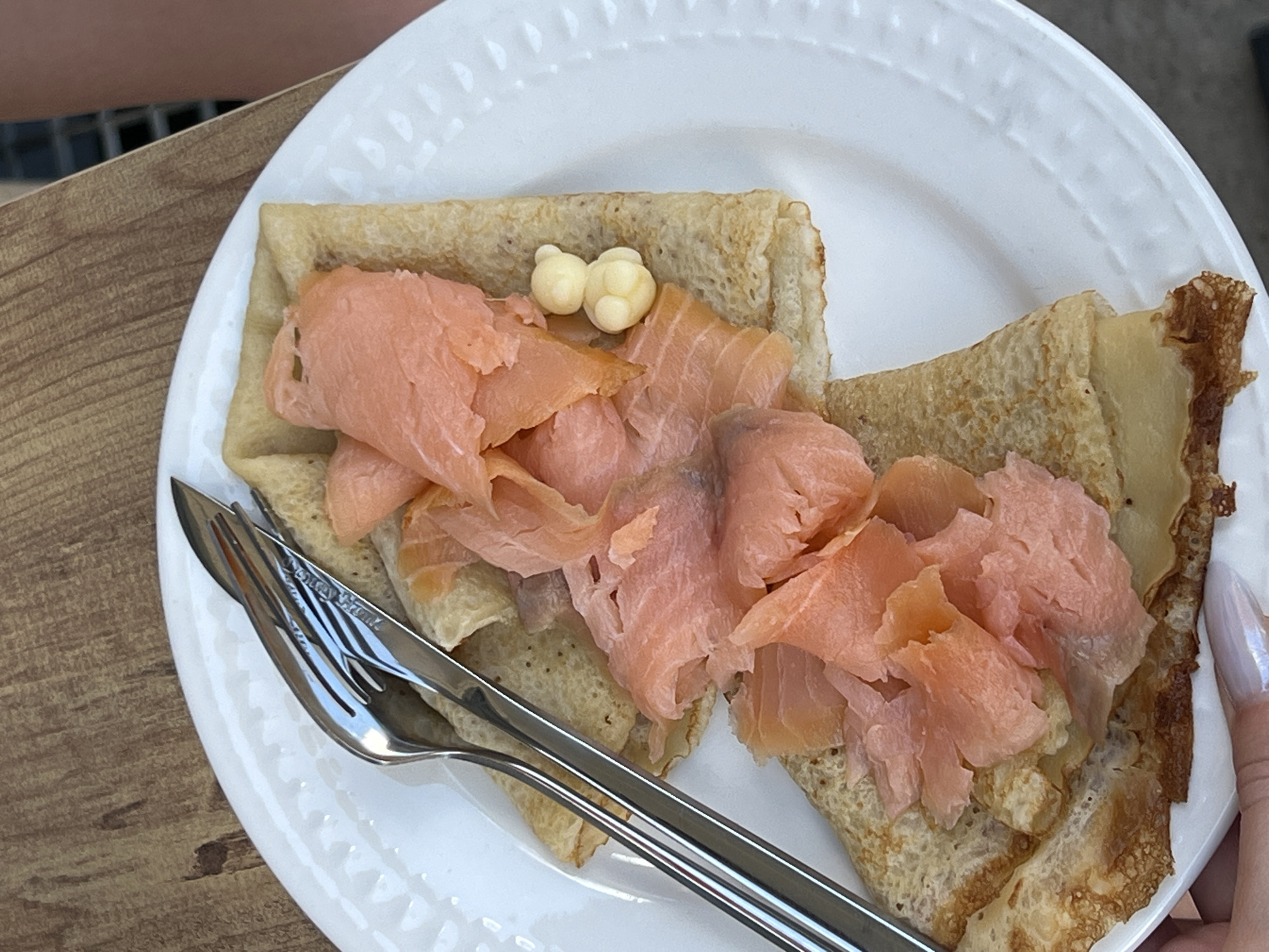 A top-down shot shows a white plate with a crepe topped with smoked salmon and two small butter balls. A silver fork and knife rest on the plate to the left of the crepe. The plate sits on a wooden table next to a concrete surface. A woman's hand with long, shiny, white nails is on the right edge of the plate. The image was taken in Budva, Montenegro.