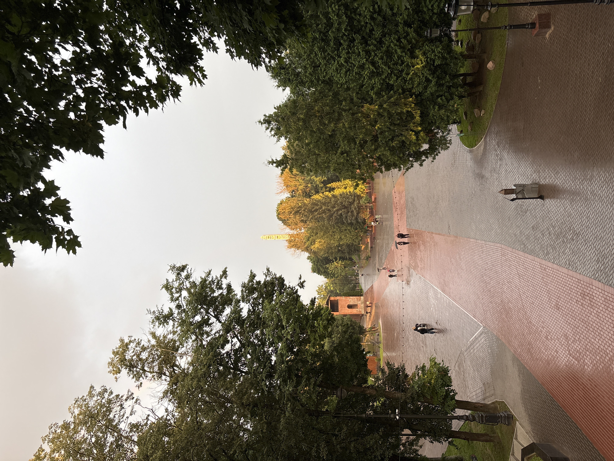 This image depicts an outdoor public space, likely a park or plaza, in Sof'ino, Russia. The ground is paved with brick and cobblestones and appears wet, suggesting it has recently rained or is currently raining. Several individuals are present, walking across the paved area, some holding umbrellas, indicating inclement weather.
On the left side of the frame, a large brick structure, possibly a tower or old building, stands prominently. To the right and in the background, various trees form a natural boundary and add greenery to the scene. Further in the distance, a tall, slender structure, potentially a radio tower or similar antenna, is visible. The sky is overcast and pale, contributing to a diffused light. The overall atmosphere is calm, with the wet surfaces reflecting the ambient light.