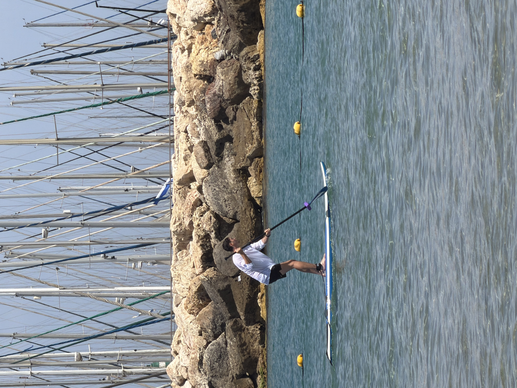 A man paddles on a stand-up paddleboard in the Mediterranean Sea off the coast of Tel Aviv, Israel. The man is wearing a white shirt and dark shorts. He is using a paddle to propel himself through the water. The coastline is composed of large rocks. Yellow buoys float in the water, possibly marking a swimming area or other boundary. In the background, a multitude of boat masts rise, indicating the presence of a marina or harbor.