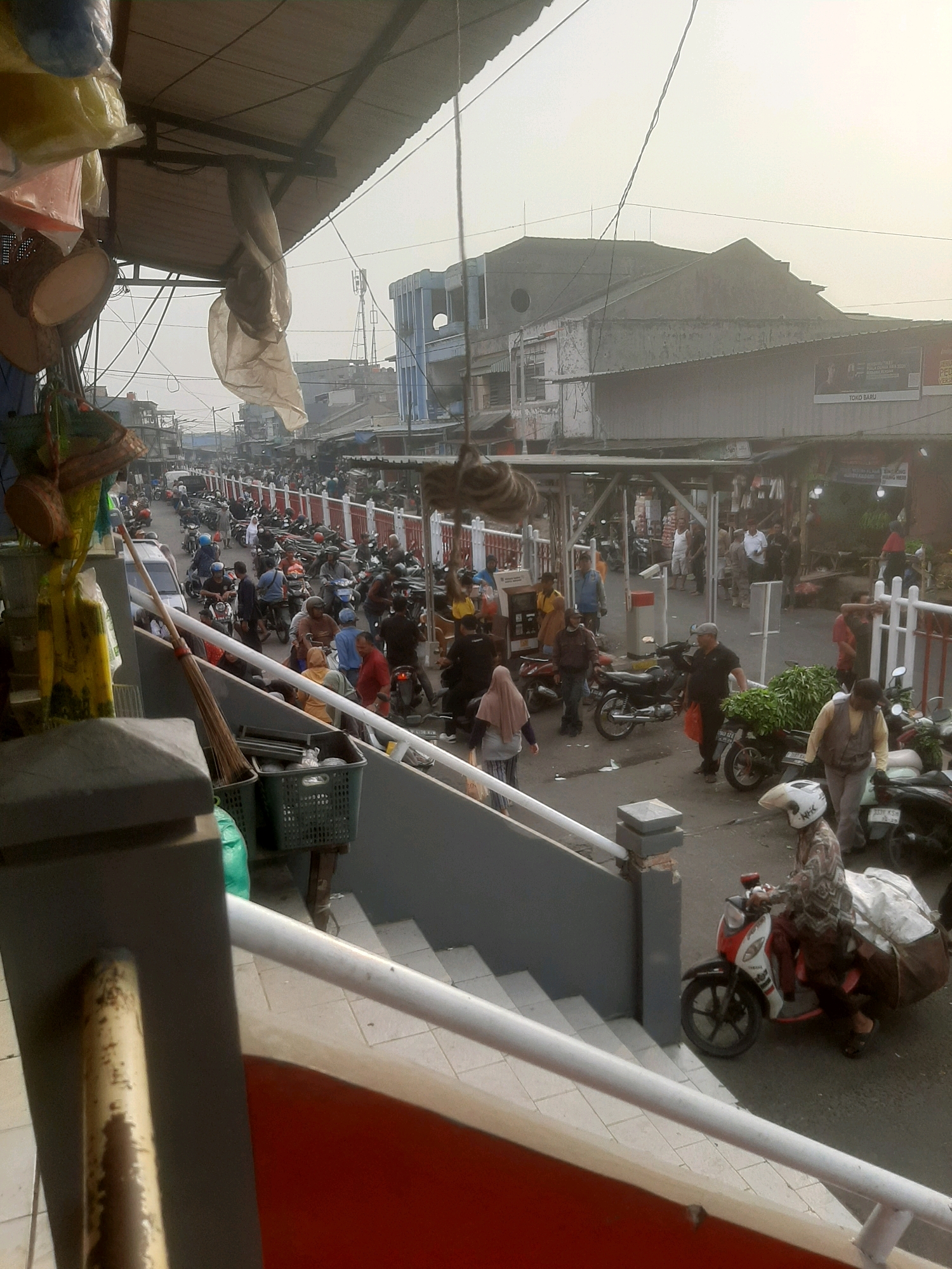 The image depicts a bustling market scene in Bekasi, Indonesia, characterized by a lively street filled with people and motorcycles. The foreground shows a weathered railing and steps, hinting at an elevated vantage point overlooking the activity.
The street is densely populated with individuals engaged in various activities. Many are seen riding motorcycles, some of which are laden with goods, indicating transportation and commerce. The people are dressed in casual attire, with some women wearing headscarves. The overall atmosphere suggests a busy marketplace with ongoing transactions and movement.
Objects in the scene include numerous parked and moving motorcycles, a white car, baskets filled with produce, and various stalls or shops lining the street. The buildings in the background appear to be typical of urban Indonesian architecture, with some showing signs of wear. The sky is overcast, contributing to a soft, diffused light. The presence of a telecommunication tower in the distance further establishes the urban setting.
The composition of the image, with its focus on the street level activity from an elevated perspective, captures the dynamic energy of the market. The scene is a snapshot of daily life in a commercial area of Bekasi.