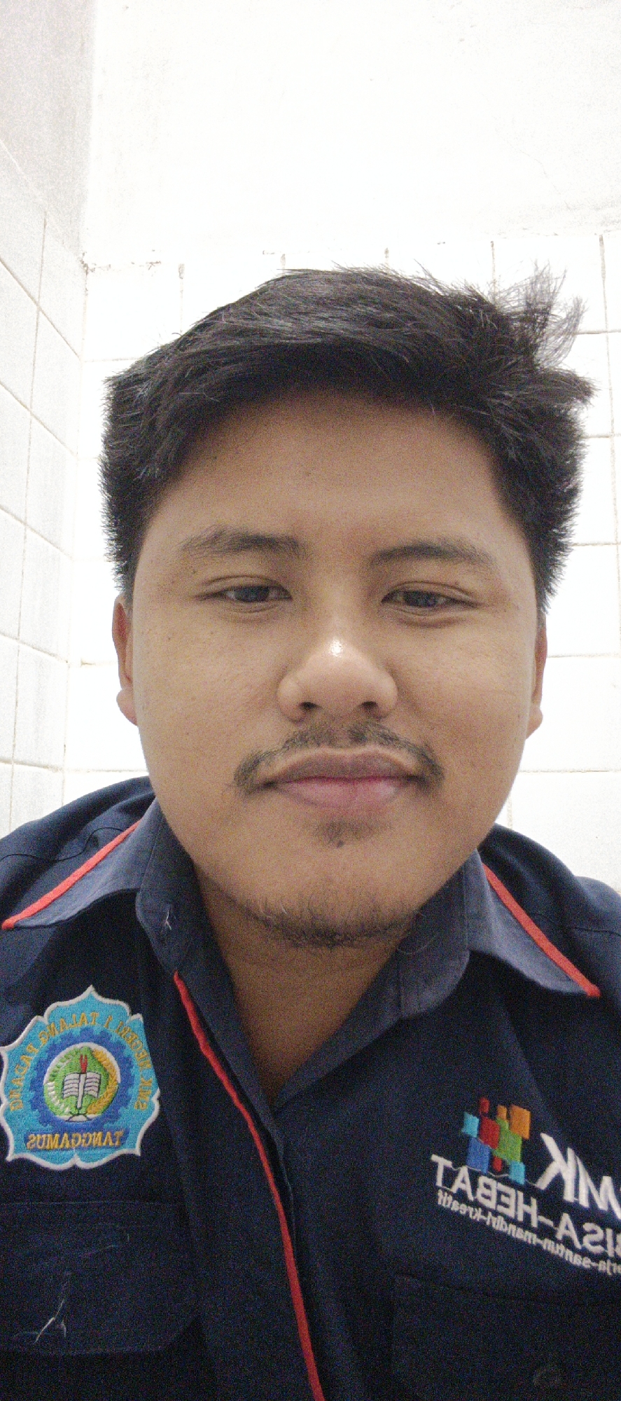 This media file is a close-up selfie of a young man. He is wearing a dark blue collared shirt with red trim and two patches. The patch on his left chest is circular and features a design with a book and greenery. The patch on his right chest is a colorful logo with text below it. He has dark, slightly messy hair, a mustache, and a small patch of goatee. The background appears to be a tiled wall. The lighting suggests an indoor setting, possibly a bathroom or similar tiled room. The man's expression is neutral, and there are no overt indications of a specific activity or event. The visible text on the right chest patch reads "NK" in large white letters and below it, in smaller text, "Tases-H-A21" which is likely a mirrored or reversed version of text. The location context is Sukabumi, Indonesia.