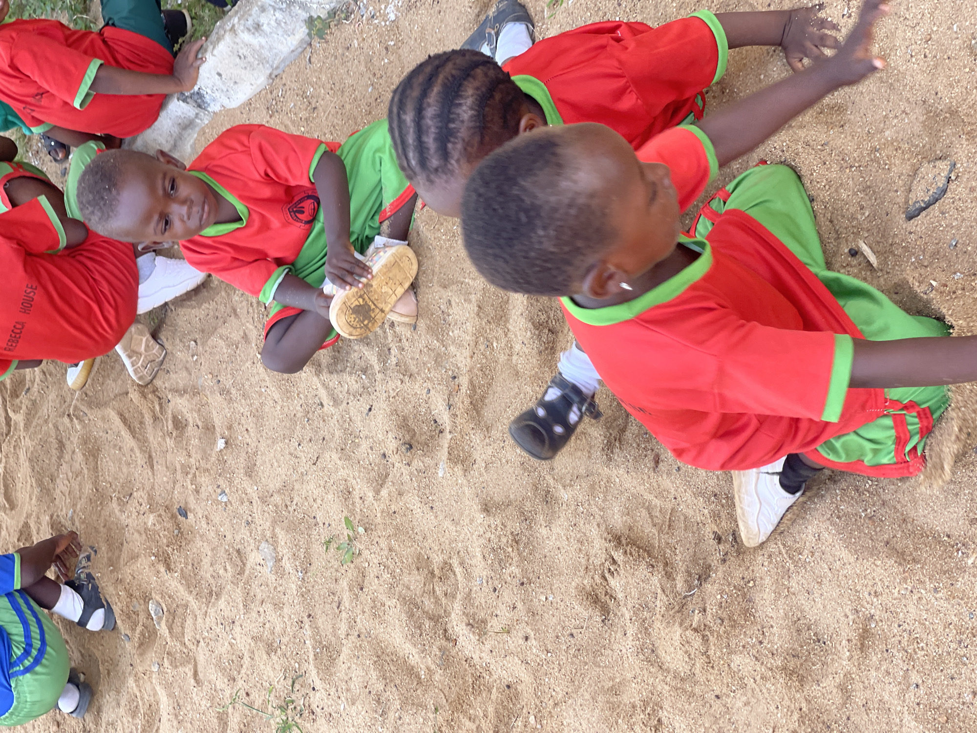 Captured from an aerial perspective, the image portrays a group of African children, mostly boys, on a sandy terrain in Akwanga, Nigeria. The children are dressed in matching red and green uniforms, likely indicating they are students. Some are seated, interacting with the sand and their shoes, while others are extending their arms, seemingly playing. The sandy area appears to be an outdoor space, possibly a schoolyard or a recreational area. In the background, there's a concrete structure. The overall setting suggests a casual, playful environment.