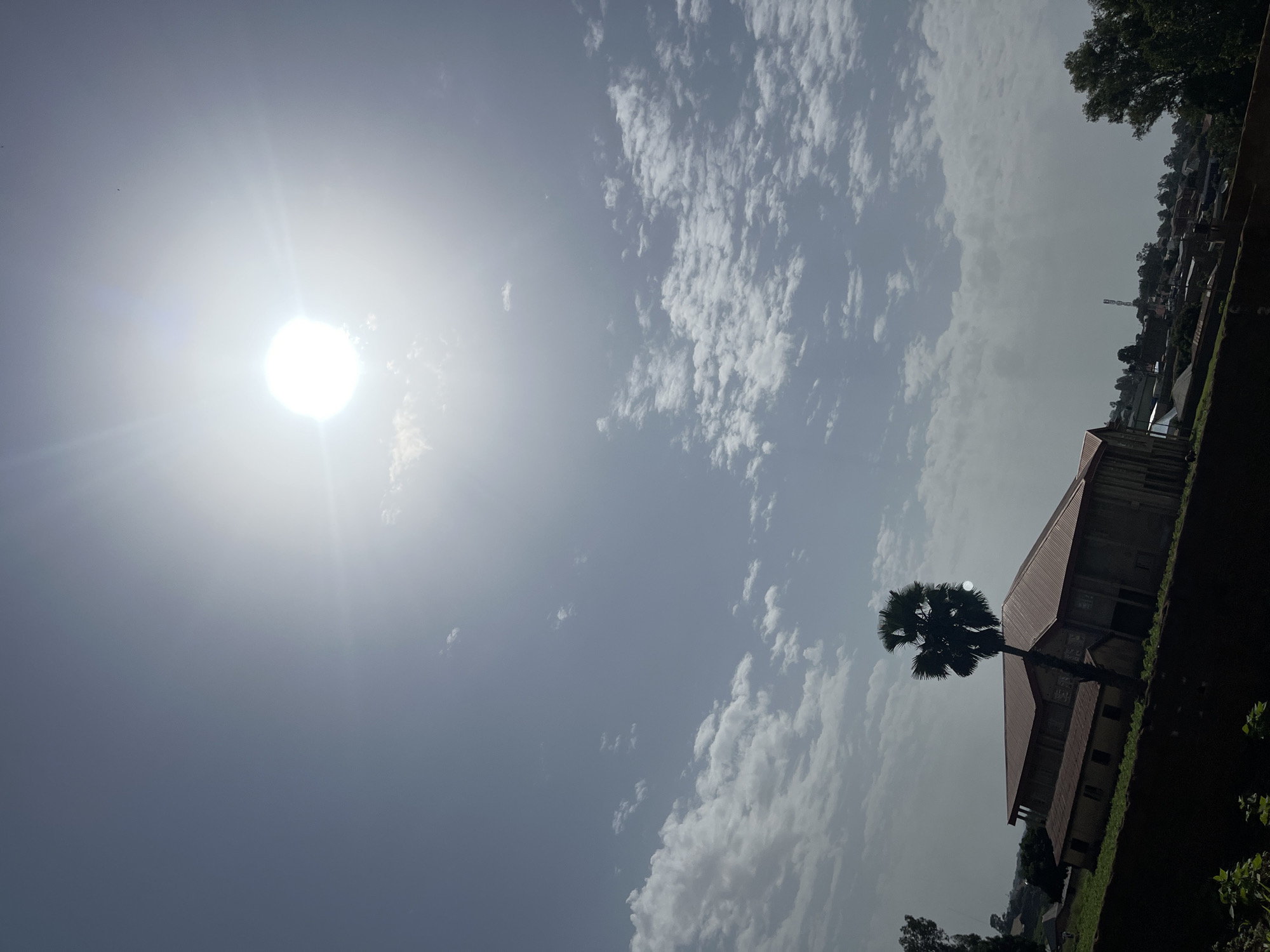The image depicts a bright, sunny day in Bukuru, Nigeria, with the sun prominently featured in the upper center of the frame, casting a strong glare. The sky is a pale blue with scattered white clouds, some appearing wispy and others more dense. In the lower half of the image, a large building with a reddish-brown roof is visible, appearing to be a school or community center, with smaller structures and trees in the background. In the foreground, a dark, sloped area suggests a retaining wall or elevated ground, with some greenery at the very bottom. A palm tree stands to the left of the building, its fronds silhouetted against the bright sky. The overall impression is of a warm, outdoor environment in a semi-urban setting.
