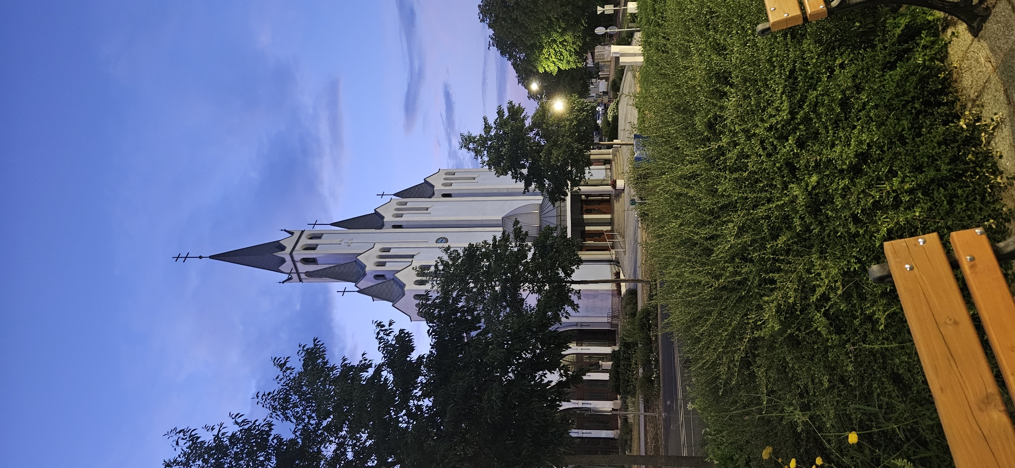 A white church with dark gray spires stands prominently against a twilight sky with clouds. The church has multiple pointed roofs and a tall central steeple topped with a double-barred cross. Arched windows and decorative elements are visible on the church facade. In the foreground, lush green bushes and trees partially obscure the view of the church entrance. Two wooden park benches with dark metal frames are positioned in the lower portion of the image, one in the foreground and another partially visible on the right. Streetlights are illuminated, casting a warm glow on the surroundings. The scene appears to be set in a public area within the city of Hévíz, Hungary, likely in front of the Church of the Holy Spirit.