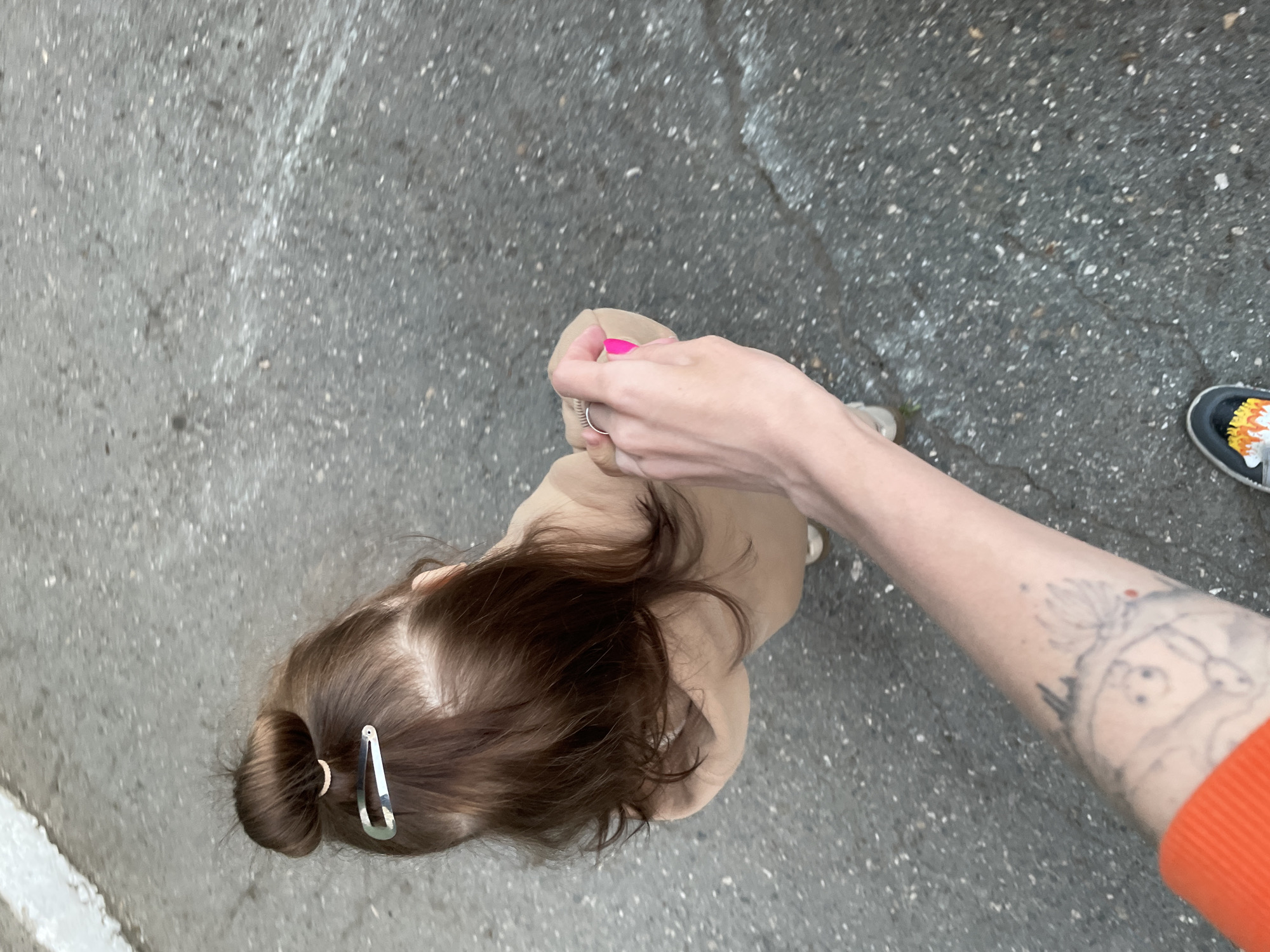 An overhead view captures two individuals on a textured, gray asphalt or concrete surface. The ground exhibits various light speckles and faint lines.
In the lower-left to central portion of the frame, the top and back of a person's head and upper body are visible. This individual has long, dark brown hair styled into a neat bun at the back, secured with a distinctive silver-colored hair clip. They are wearing a light-colored, possibly beige or light brown, garment.
Extending from the right side of the frame, an adult's arm and hand are prominent. The arm is clad in an orange ribbed sleeve and displays a tattoo on the forearm, featuring a detailed, possibly cartoon-like or stylized design with a small red dot near the top. The hand has bright pink nail polish and wears a silver ring on one finger, appearing to gently hold or interact with the garment of the first person. A black shoe with vibrant yellow, orange, and white accents is partially visible in the upper right corner, belonging to the adult.
The image suggests an outdoor moment, possibly a casual walk or interaction, in Izhevsk, Russia. No discernible text is present in the scene.