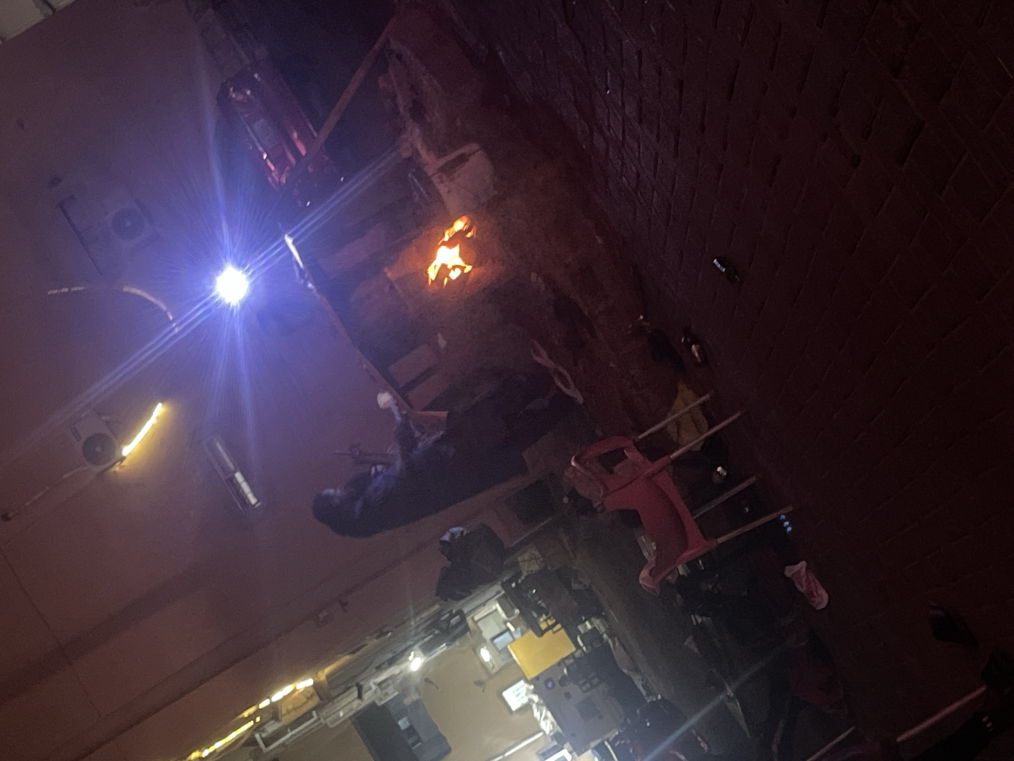 The image, taken in Lugbe, Nigeria, shows a street scene at night. A bright light source illuminates the area, causing lens flare. In the middle ground, there appears to be a small fire. Several people are present, though their details are obscured by shadows and the low light. Various objects, including what appears to be a red chair, are scattered around the street. The environment includes brick walls on one side and buildings on the other.
