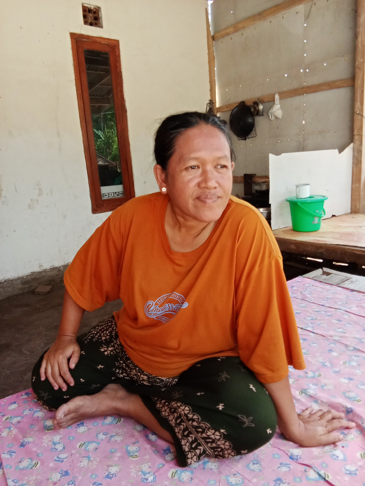A medium shot captures a woman of East Asian descent sitting cross-legged on a pink patterned blanket on the floor. The woman has dark hair pulled back, a round face, and is wearing an orange t-shirt and dark green patterned pants. She is looking to her right, with her left arm resting on her knee and her right hand extended forward.
The environment appears to be a simple outdoor or semi-outdoor structure. To the left of the woman, a cream-colored wall has a narrow, vertically oriented wooden window with a reflective pane. Above the window is a small square vent. To the right, the background consists of unfinished wooden beams and walls, with a gray paneled wall where kitchen items are visible, including a black pot and a white cloth hanging. Further to the right, on a wooden surface, sits a bright green plastic bucket with a white cup on top.
The floor in front of the woman is made of concrete, and the blanket she is sitting on has a repeating pattern of cartoon characters, likely Hello Kitty. The scene is well-lit by natural daylight, suggesting it is daytime. The overall impression is a humble, domestic setting in what could be a rural area of Selowogo, Indonesia.