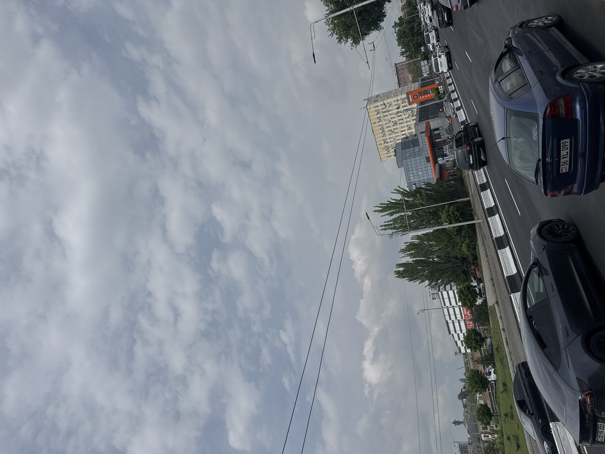 The image depicts a street scene in Yerevan, Armenia. The view is of a busy road with multiple lanes filled with cars. Buildings and trees line the street. The sky is overcast with clouds. A large "SAS" supermarket sign is visible. The cars include a blue Opel Astra with license plate "36 XL 099" and a black Tesla with license plate "55 HA 055".