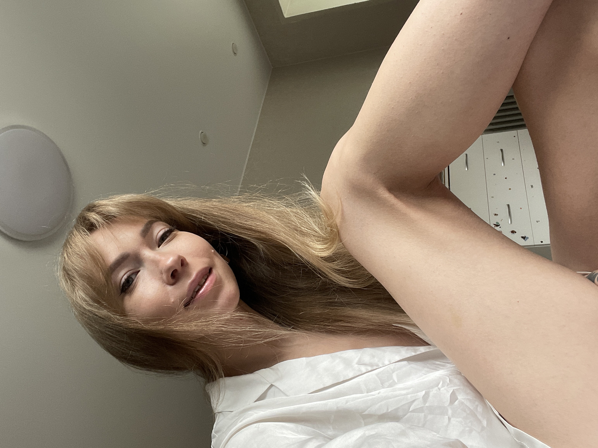 The image shows a young woman with blonde hair and bangs, wearing a white collared shirt. She is sitting, and her legs are crossed. The background is an indoor setting, possibly a bedroom, with a dresser visible. There is a light fixture on the ceiling. The image does not contain any elements that indicate a specific location in the city of Perm, Russia.