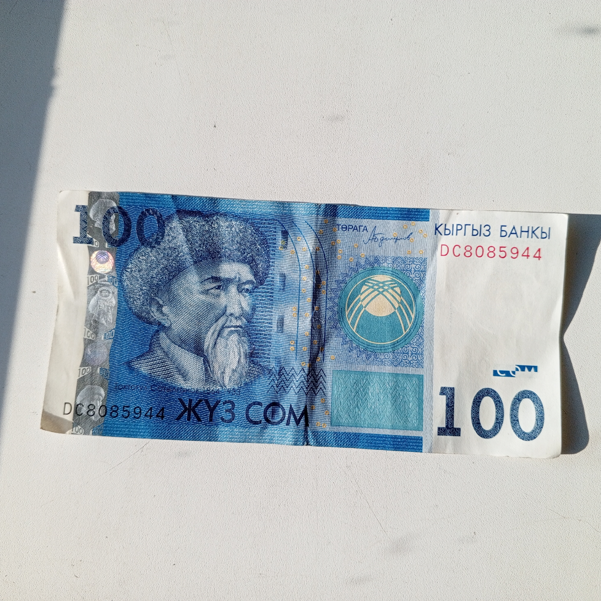 The image shows a close-up, top-down view of a single 100 Kyrgyzstani Som banknote lying on a white, textured surface. The banknote is partially illuminated by a shadow from the left, creating a diagonal line across the upper portion of the image. The banknote features a portrait of a man with a beard and a fur hat on the left side, and the number "100" prominently displayed on the right. Text in Cyrillic script is visible, including "КЫРГЫЗ БАНКЫ" (Kyrgyz Bank) and a serial number "DC8085944". The banknote is oriented horizontally. There is no indication of the city of Sayanogorsk, Russia, or any urban environment in the image.