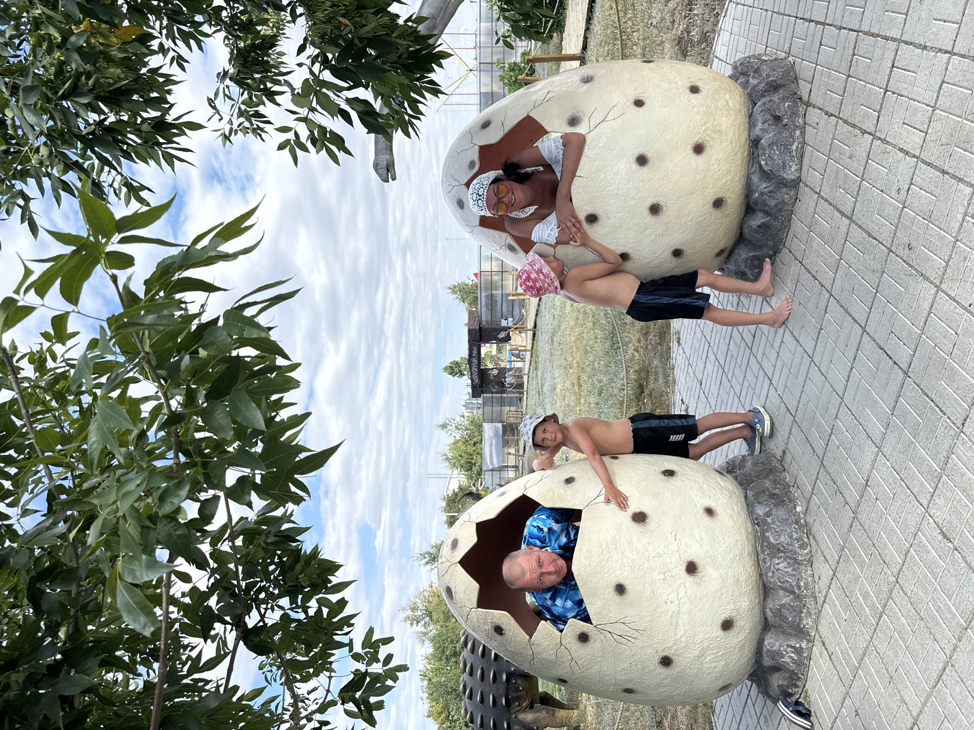 An adult male and an adult female are emerging from large, cracked egg-shaped sculptures, with two young boys standing near them. The male, wearing a blue patterned shirt, leans out of the left egg. The female, in a white top and a patterned headscarf, smiles from the right egg. One boy, wearing a pink hat and black shorts, stands near the female. Another boy, in a grey hat and black shorts, is by the male. They are in an outdoor park with a paved walkway, grass, trees, and dinosaur-like sculptures in the background, suggesting a dinosaur-themed attraction. The sky is partly cloudy. The scene is located in the city of Sol-Iletsk, Russia, at the "DinoMir" dinosaur park.