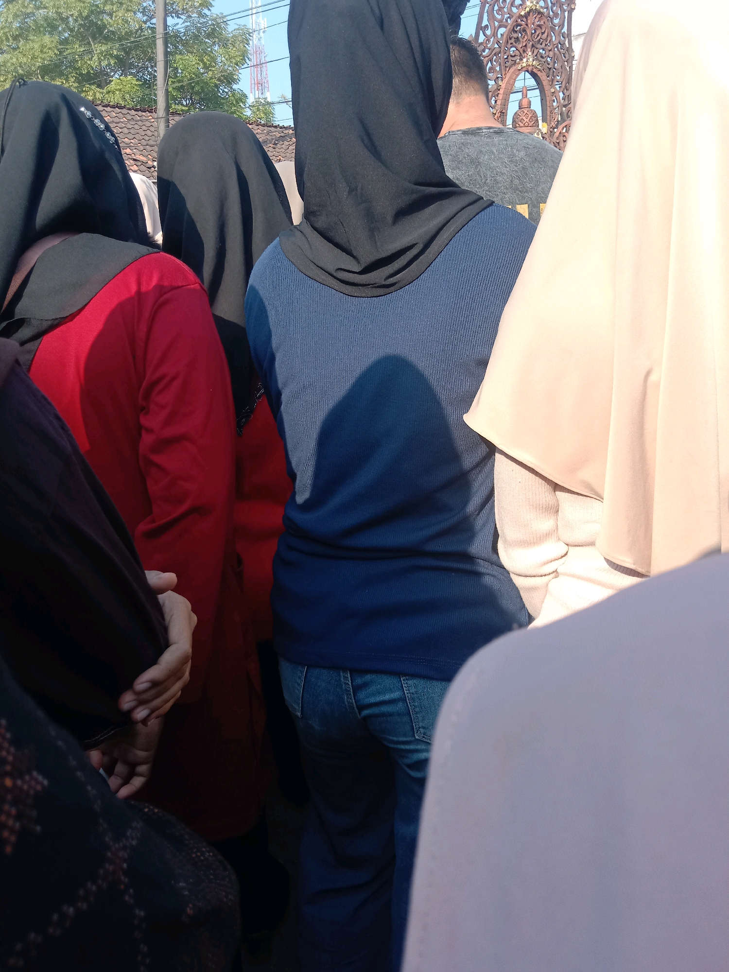 In Banjardowo, Indonesia, a group of people, predominantly women wearing hijabs, stand outdoors under a clear sky. The sunlight casts shadows on the ground. In the foreground, the backs of several individuals are visible. A woman on the left wears a red long-sleeved shirt, while the woman in the center wears a blue long-sleeved ribbed shirt and blue jeans. To the right, a person is partially visible wearing a light-colored hijab and sweater. Behind them, a man with short hair wearing a gray t-shirt can be seen. The background features trees with green foliage, a tiled roof, and a tall tower with antennae. Decorative metalwork is visible in the upper right portion of the frame. One person's hand, with manicured nails, is held near their chest in the lower left.