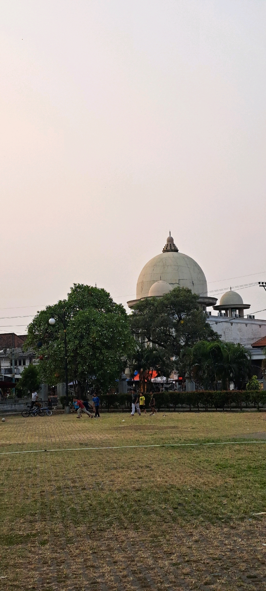The image depicts an outdoor scene in Bangil, Indonesia, featuring a large park area in the foreground and a building with a prominent dome in the background. The park has a grassy field, partially mowed with some patches of bare soil, and a white line marking the edge of the field. Several people are present in the park. In the middle ground, a group of young men and boys are engaged in activities, including what appears to be a game of soccer. One individual is kicking a soccer ball, while others are running and positioned on the field. Two bicycles are parked on the left side of the image.
Behind the park, a large, light-colored building with a distinctive dome and smaller domed structures is visible. The building appears to be a place of worship or a significant civic structure. Trees, including a large, leafy tree with a street lamp next to it, stand between the park and the building. The sky is a pale, overcast hue, suggesting it may be dawn, dusk, or a heavily clouded day. The overall impression is a tranquil public space in the late afternoon or early morning.