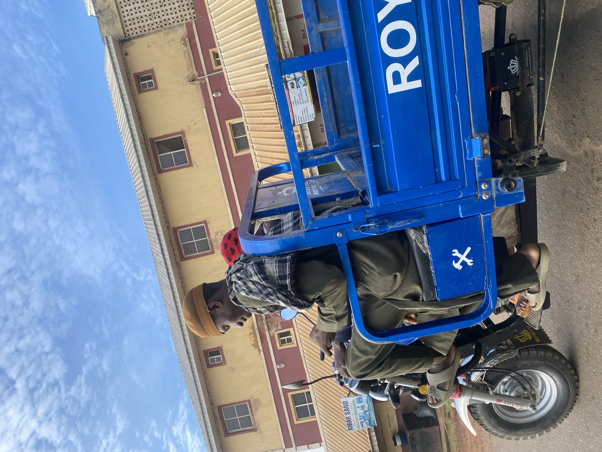 In the city of Rafin Par, Nigeria, a three-wheeled vehicle, painted royal blue, is captured from a low angle, moving along a paved road. The vehicle is laden with what appears to be cargo in its rear bed. Two individuals are seated on the vehicle. The driver, positioned in the front seat, is a young man wearing a brown knitted cap, a green robe, and a checkered scarf draped over his shoulders. He is looking down at a black mobile device held in his hand. A second individual, partially visible behind the driver, wears a red cap with white polka dots. The building in the background is a two-story structure with yellow-painted walls and a maroon trim. Its windows are framed in a dark color, and the roof is covered with corrugated metal. The sky above is a bright blue with scattered white clouds. A sign advertising "Amar Sano Business Center" is visible on a pole near the building. Another sign on the blue vehicle, partially obscured, reads "ROYAL MOTORCYCLE".
