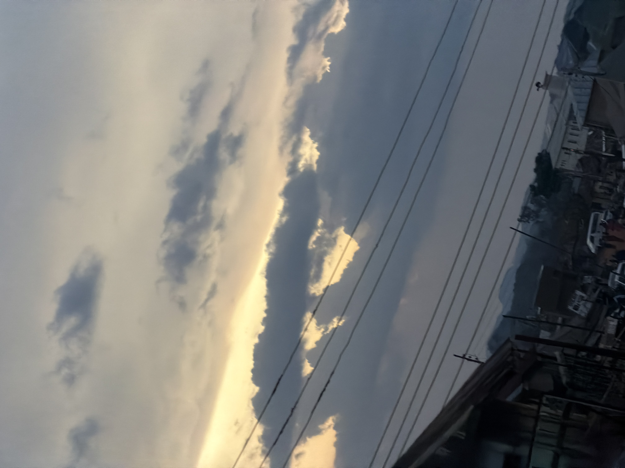 This is a low-angle, vertical, medium shot of a cloudy sky over a dense urban environment, likely in Bukuru, Nigeria. The sky dominates the upper two-thirds of the frame, with layers of grey and white clouds, some illuminated by a soft yellow glow, suggesting either sunrise or sunset. Multiple power lines stretch across the sky, obscuring parts of the cloud formations.
The lower third of the image shows a portion of a settlement. On the left, a dilapidated building with a corrugated metal roof is visible, partially obstructing the view of what appears to be a street scene. In the background, several buildings, some with light-colored facades, are scattered amongst trees. A few vehicles, including what looks like a white van, are present on the ground. Distant hills or mountains are faintly visible against the hazy sky. The overall impression is one of a densely populated area under a dramatic sky.