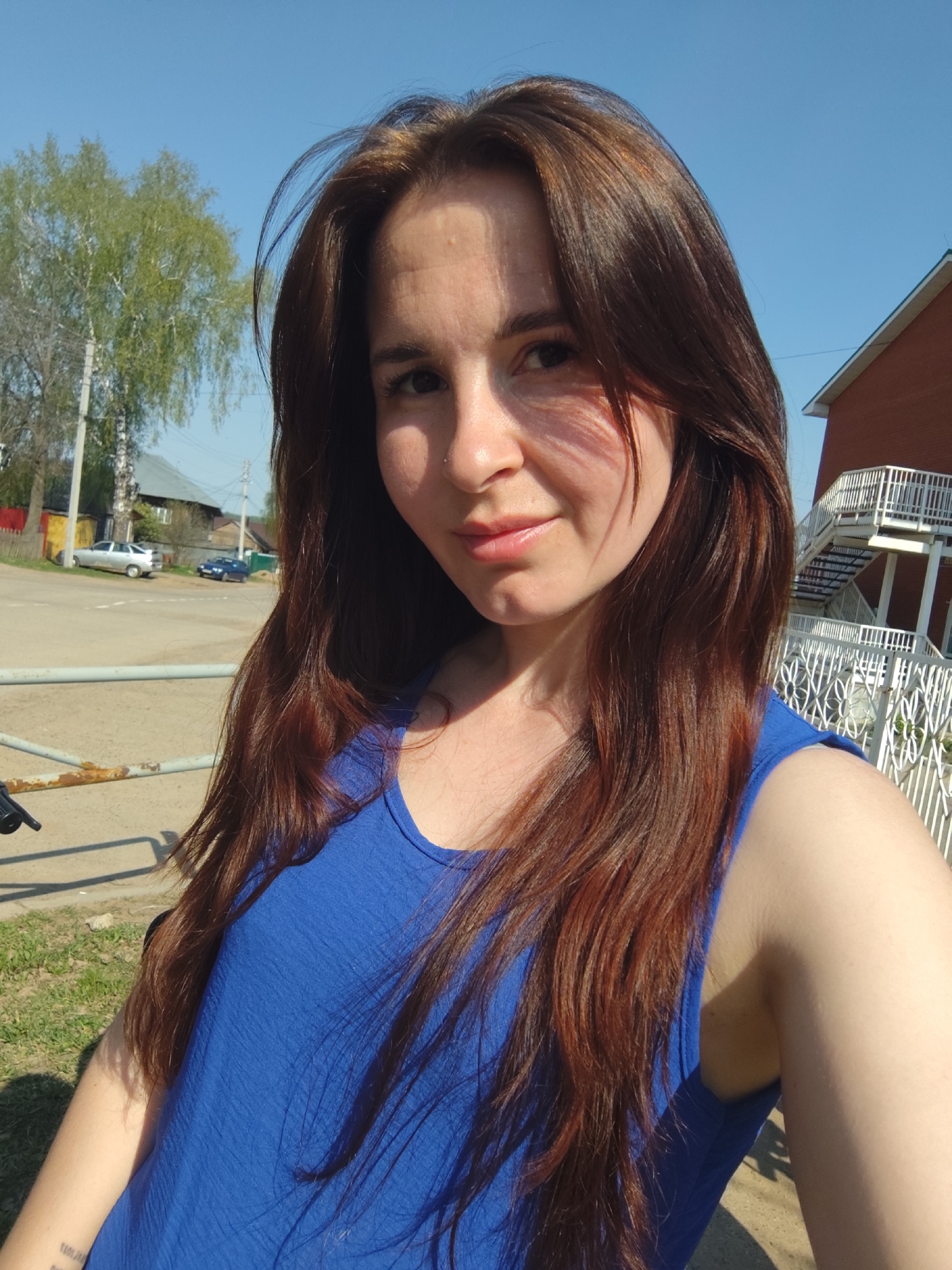 A woman is taking a selfie outdoors on a sunny day. She has long, reddish-brown hair and is wearing a blue top. A nostril piercing is visible on her left nostril. The background includes trees, a road with cars, a fence, and a brick building with a white exterior staircase. The location appears to be in a residential area. The location is likely Фоки, Russia.