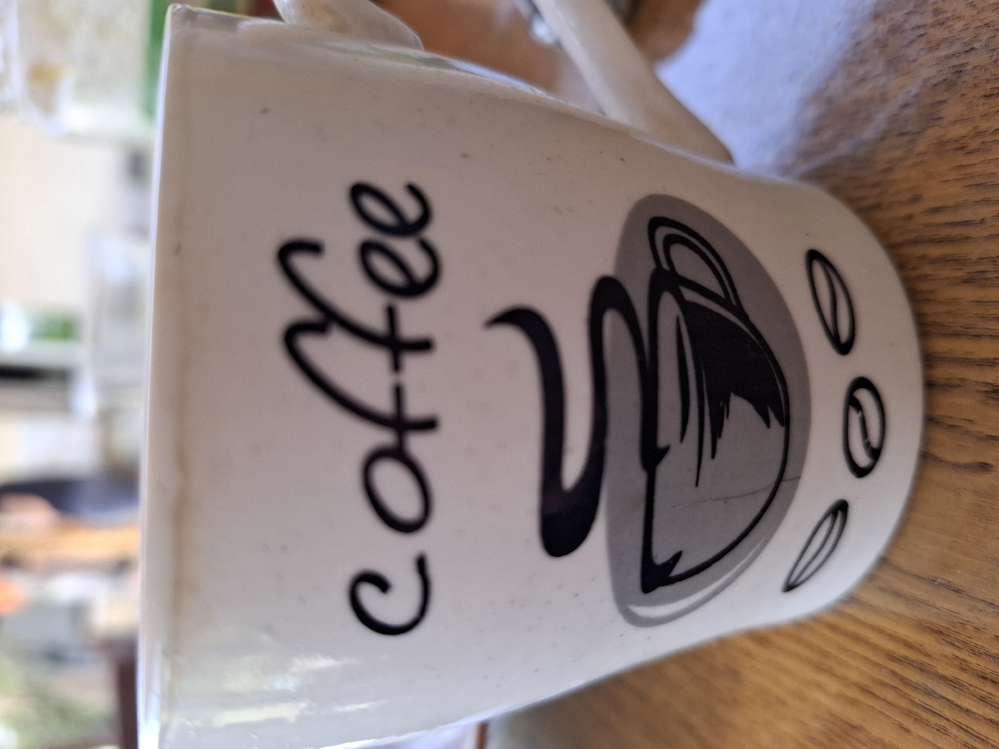 The image shows a white coffee mug with the word "coffee" written in black cursive script. Below the word is a graphic of a steaming coffee cup. There are also two coffee bean shapes depicted near the bottom of the mug. The mug is sitting on a wooden surface, likely a table. The background is blurred, showing a person in the distance and some bright, out-of-focus objects. No specific location in the city of Galabovo, Bulgaria can be identified from the image.