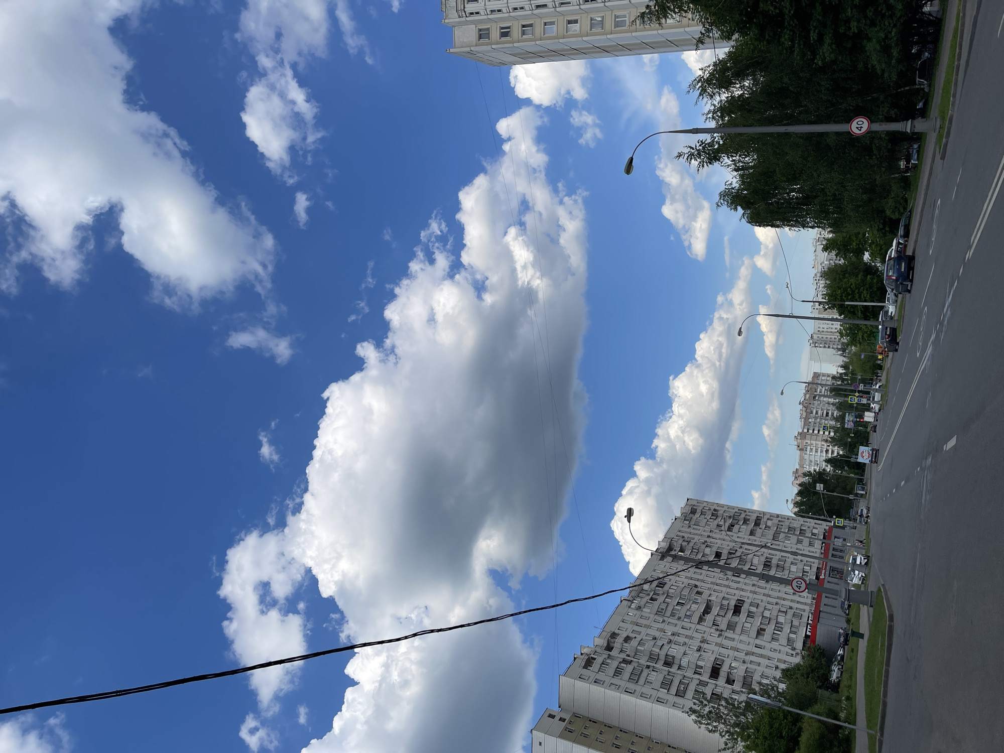 This media file captures an urban street scene in Moscow on a bright, sunny day. The dominant feature is a vibrant blue sky filled with numerous fluffy white cumulus clouds, indicating fair weather.
The scene includes tall, multi-story residential buildings, typical of Soviet-era panel construction, which line the edges of a wide asphalt road. One prominent grey building, with many windows and balconies, is seen on the lower right, its base featuring a red commercial facade with indistinct text. Another similar building is visible further in the distance on the left side of the road.
A multi-lane road runs horizontally across the lower part of the image, with parked cars visible along its side. Lush green trees line the sidewalk, separating the buildings from the street. Several dark streetlights with curved tops are spaced along the road, with power lines crisscrossing the sky in various parts of the frame.
A circular white traffic sign with a red border, clearly displaying the number "40" in black, is attached to one of the streetlight poles, indicating a speed limit of 40 km/h. The bright lighting and shadows suggest it is midday or early afternoon. No specific human activity is visible, but the presence of vehicles and buildings implies a typical urban environment.