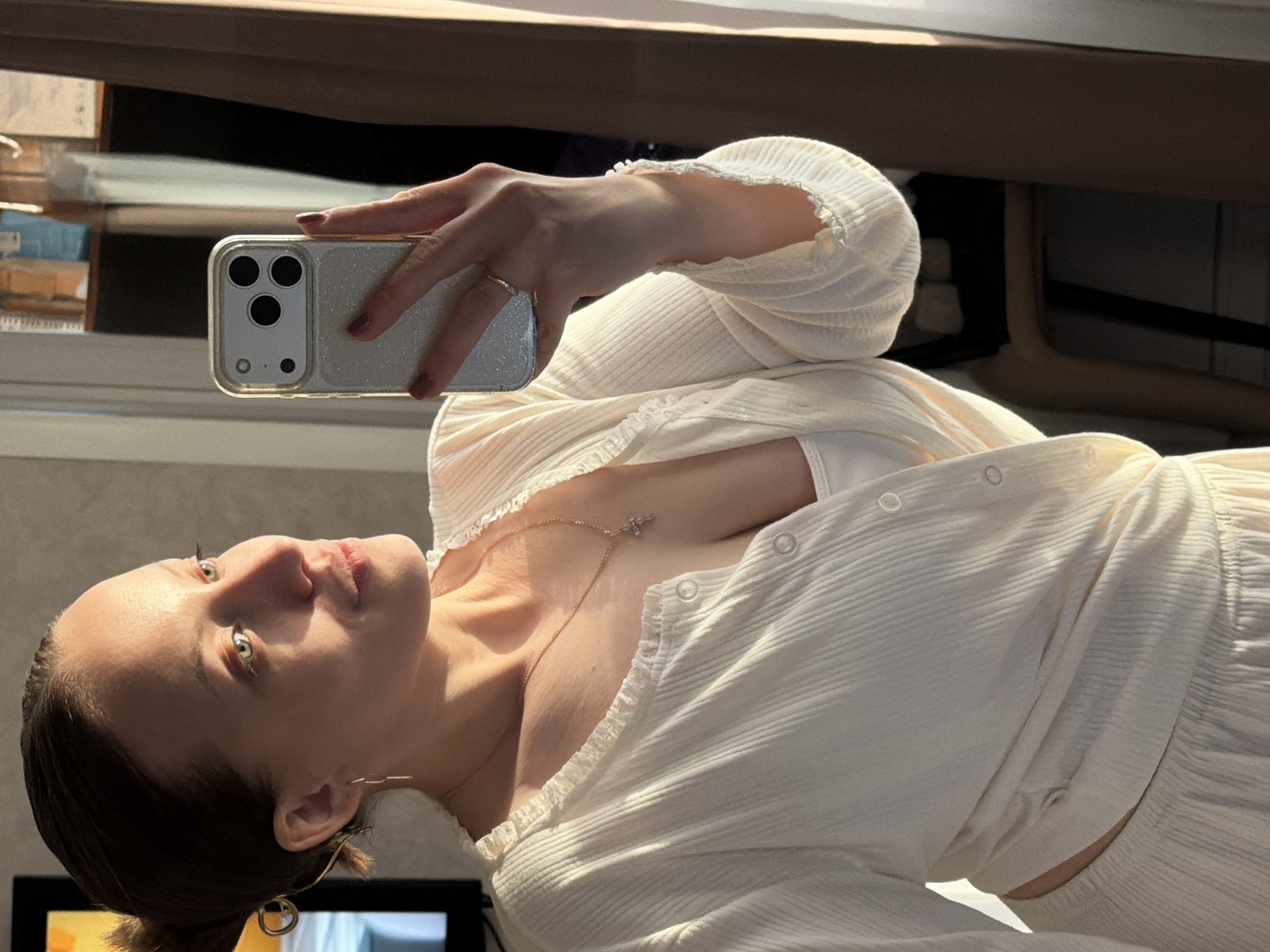 A woman is taking a selfie with a silver iPhone. She is wearing a white top and has a necklace. The background includes a window and furniture. The location is not discernible as Reshetnikovo, Russia.