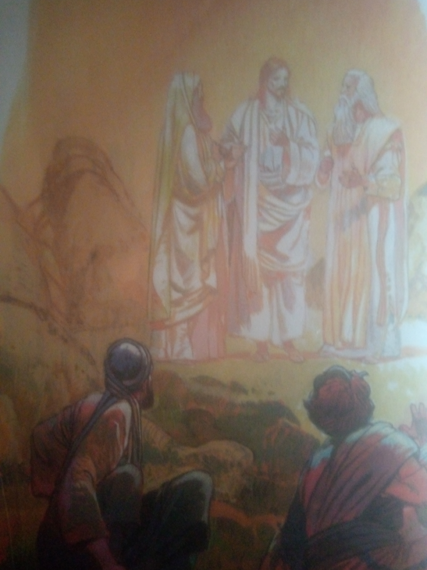 The image depicts a group of figures in a landscape that appears to be a desert or arid region. In the foreground, two individuals are seated, facing away from the viewer and towards a central group of three standing figures. The standing figures are dressed in flowing robes, reminiscent of historical or religious attire. One figure in the center appears to be the focal point, with the other two figures on either side. The background features stylized rock formations or hills. The overall color palette is warm, dominated by earthy tones, oranges, and yellows, suggesting a sunny or desert environment. The image has a soft, slightly blurred quality, as if it is an illustration or a painting. There is no identifiable element within the image that specifically places the scene in the city of Jauro Yinu, Nigeria, or any other specific urban or geographical location.