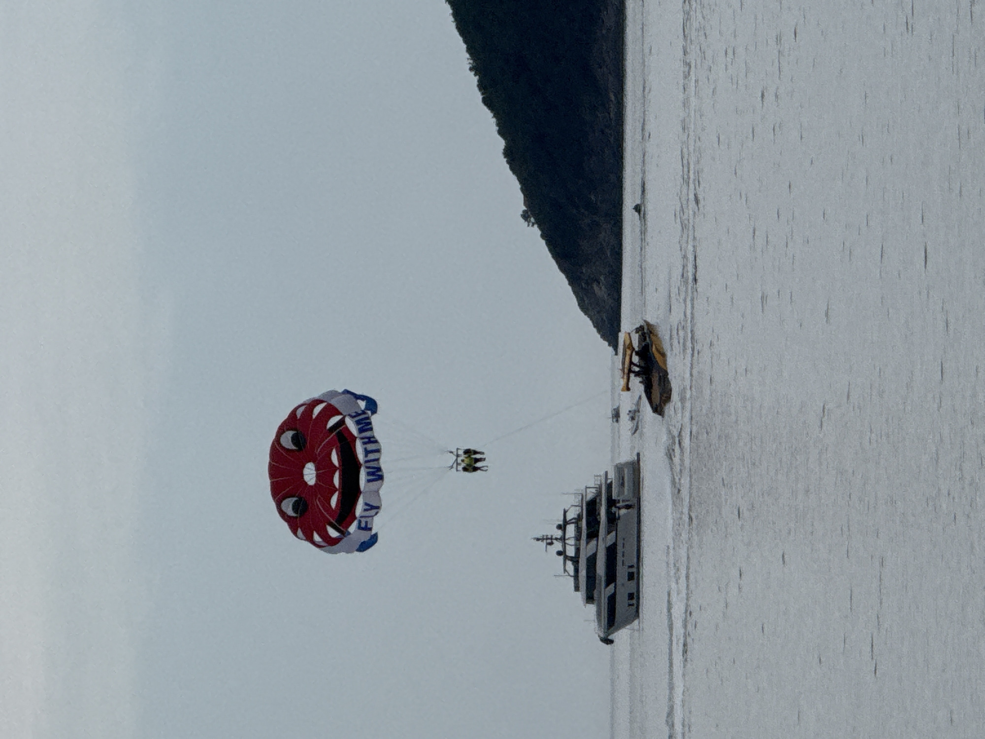 The image shows a parasailing scene over the Adriatic Sea, likely near Budva, Montenegro, given the mountainous coastline in the background. A parasail with a red and white smiling face design and the words "FLY WITH ME" in blue is in the air, suspended by ropes. Two people are visible beneath the parasail, presumably the parasailers. A large, multi-tiered boat is below the parasailers on the water. A smaller yellow boat is nearby. The sky is overcast, and the water surface appears slightly choppy.