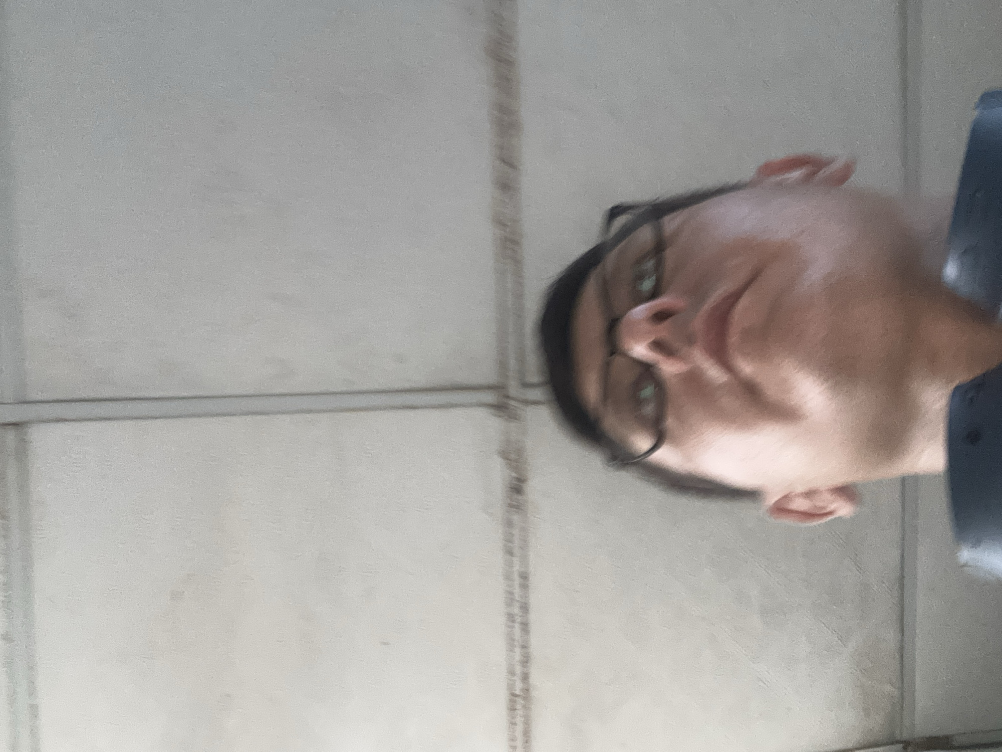 A low-angle, close-up, blurry, head-and-shoulders shot of a person wearing glasses is captured from a perspective looking up at them, suggesting the photo was taken from a floor or lower position. The individual's face is partially obscured, with their glasses framing their eyes and their skin tone appearing fair. The background consists of a textured, light-colored ceiling with visible seams and subtle discoloration, indicating an indoor environment, possibly a room or hallway with fluorescent lighting. The overall image quality is compromised by blurriness, which obscures fine details.