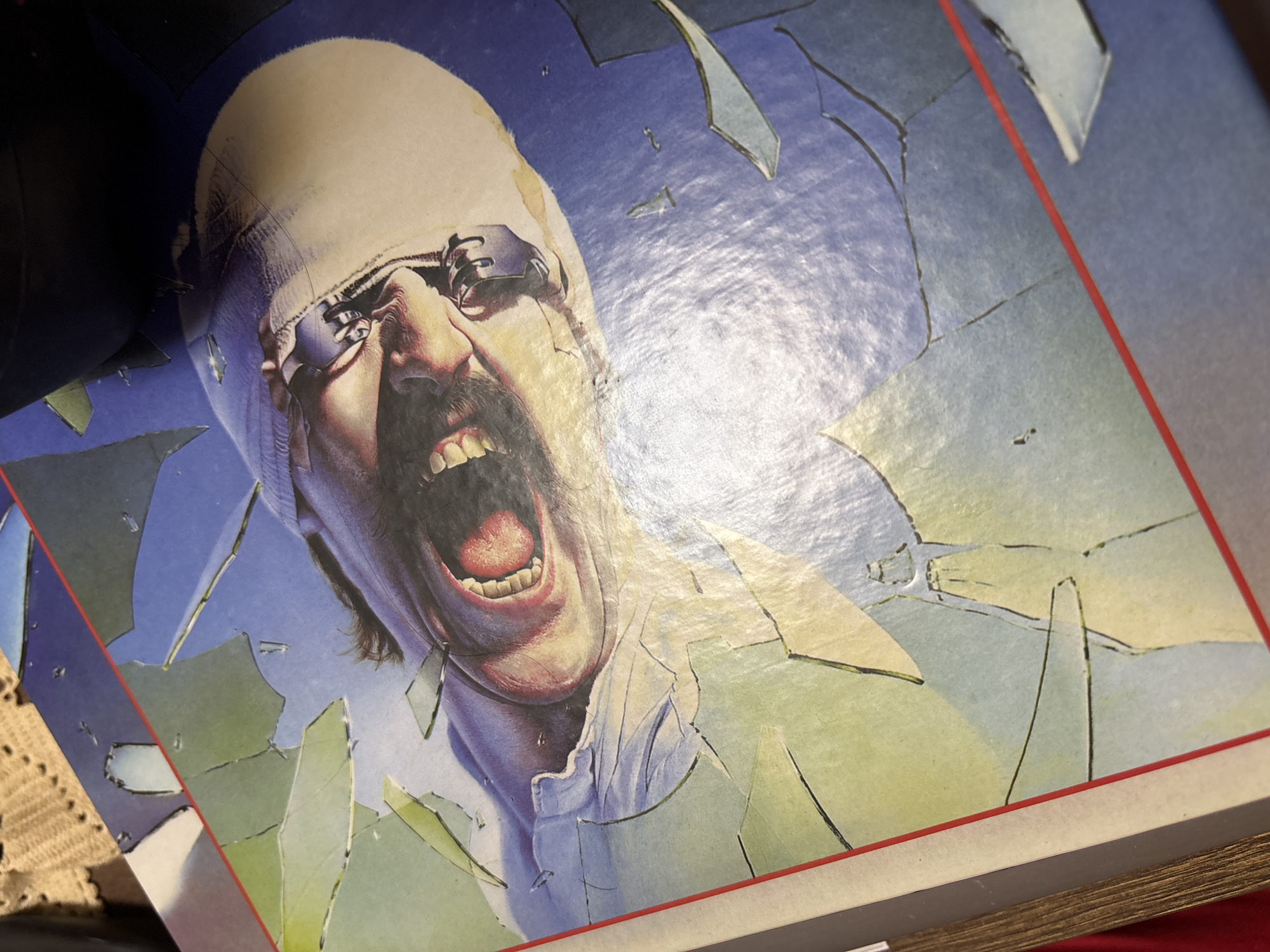 The image depicts an album cover featuring a man with a bandaged head, goggles, and a mustache, screaming. The background appears to be fractured, resembling shattered glass. The album cover is situated on a surface, possibly a table, with a crocheted object partially visible in the lower left corner. The image does not contain information to determine a location in the city of Kuklen, Bulgaria.