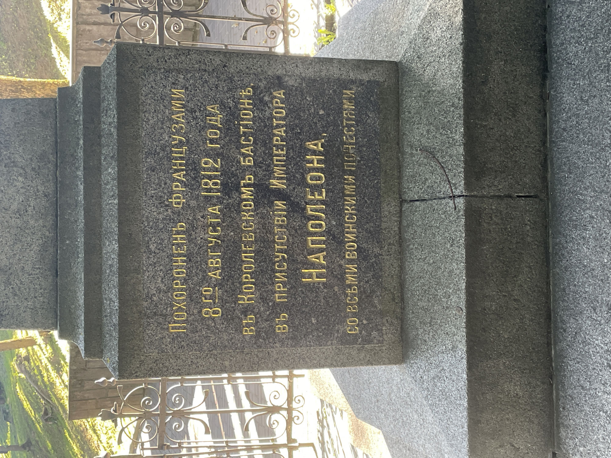 The image shows a stone monument with golden Cyrillic inscriptions. The inscription reads: "ПОХОРОНЕНЬ ФРАНЦУЗАМИ 8-ГО АВГУСТА 1812 ГОДА ВЪ КОРОЛЕВСКОМЪ БАСТІОНЪ, ВЪ ПРИСУТСТВІИ ИМПЕРАТОРА НАПОЛЕОНА, СО ВСЪМИ ВОИНСКИМИ ПОЧЕСТЯМИ." This translates to: "BURIED BY FRENCHMEN ON AUGUST 8, 1812, AT THE ROYAL BASTION, IN THE PRESENCE OF EMPEROR NAPOLEON, WITH ALL MILITARY HONORS." The monument is made of granite and has a stepped base. It is located outdoors, with sunlight casting shadows on the grass and trees in the background. A decorative metal fence is visible to the left and right of the monument. The overall scene suggests a historical memorial site.