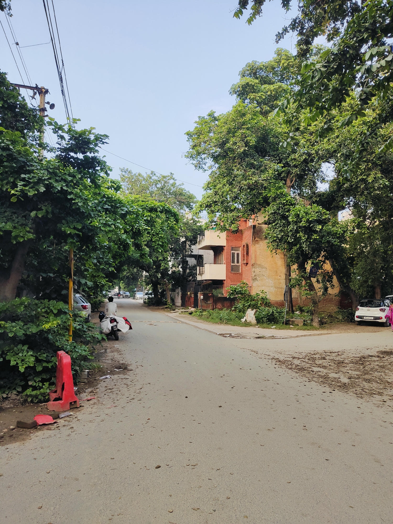 The image depicts a quiet street in Gurgaon, India, lined with mature trees and residential buildings. The road surface is asphalt, showing signs of wear and scattered debris. On the left side of the road, a person is seated on a parked scooter, which is loaded with white bags. A red traffic barrier lies on its side near the scooter. Further down the street, several cars are parked and in motion, indicating a residential area with some vehicular traffic. The buildings are a mix of styles, with one prominent red brick structure with cream-colored balconies. The sky is clear and light blue, suggesting daytime. Lush green foliage from the trees dominates the scene, creating a shaded and somewhat overgrown atmosphere. The overall impression is a typical urban street in a residential neighborhood of Gurgaon.