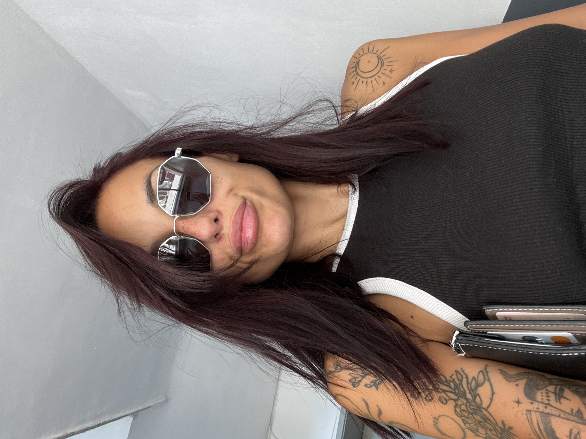 A medium shot captures a young woman from the shoulders up, facing the camera and slightly angled to her left. She has dark, layered hair, some of which is falling across her face. She is wearing large, octagonal sunglasses with a silver frame and dark lenses that reflect the environment, showing a glimpse of a street with cars. Her lips are slightly pursed. She has freckles across her nose and cheeks. The woman is wearing a black ribbed tank top with white trim around the neckline and armholes. She has tattoos on her left arm and shoulder, including a floral design on her upper arm and a sun and moon design on her right shoulder. She is holding a black wallet with cards visible in her right hand, tucked into the front of her top. The background is a plain, textured wall that appears to be indoors, with a ceiling visible in the upper left corner. The lighting is bright, suggesting daytime. There is no discernible information in the image to indicate the location in the city of Pleven, Bulgaria.