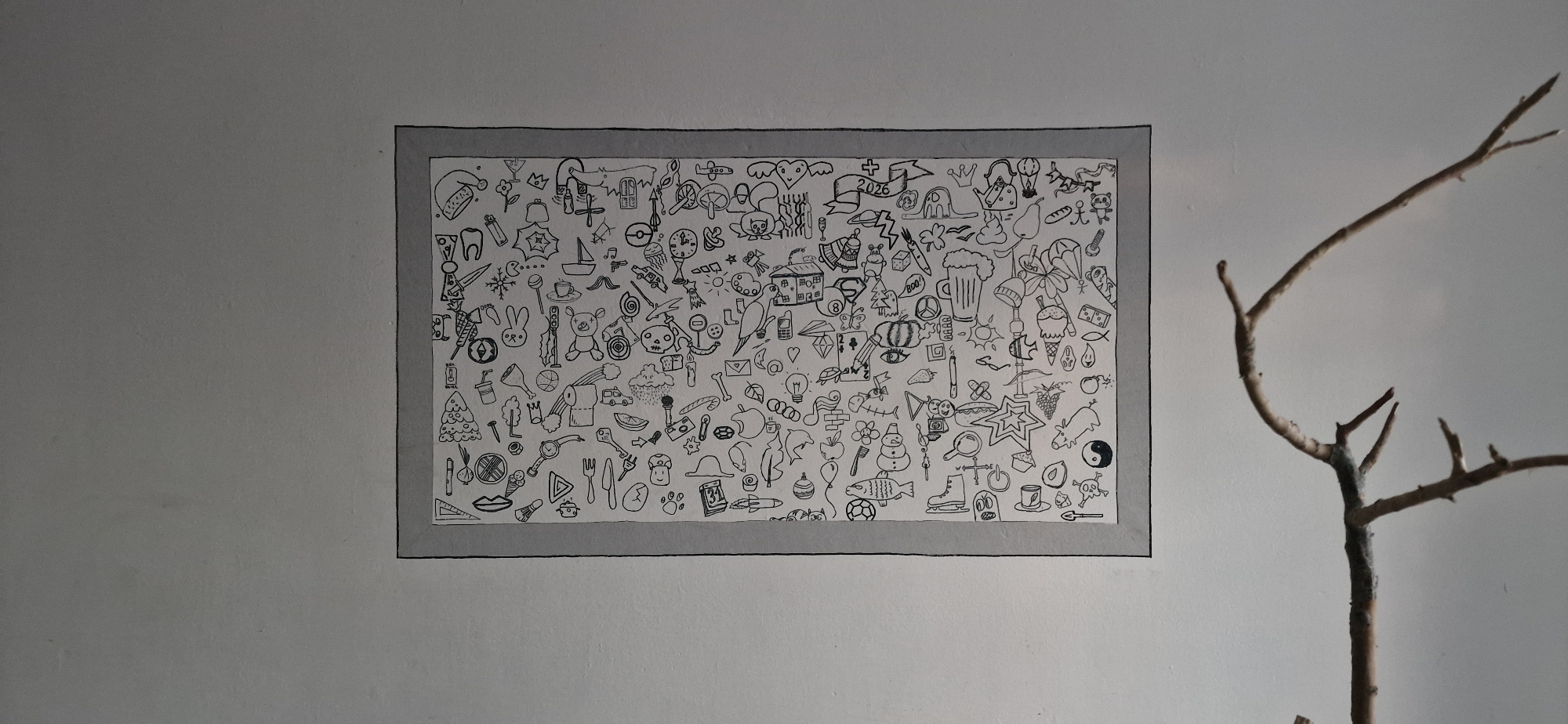 The image shows a framed drawing on a white wall. The drawing is a collage of small, hand-drawn black and white illustrations of various objects and symbols, including a tooth, a snowflake, a sailboat, ice cream cones, a teddy bear, a house, a beer mug, a Christmas tree, a snowman, a fish, a star, and a Yin and Yang symbol. To the right of the drawing, a thin, bare tree branch extends from the bottom right corner upwards. The wall appears to be a textured, painted surface. There are no people or animals visible in the image. The location of this drawing in the city of Usharal, Kazakhstan is not identifiable from the provided image.