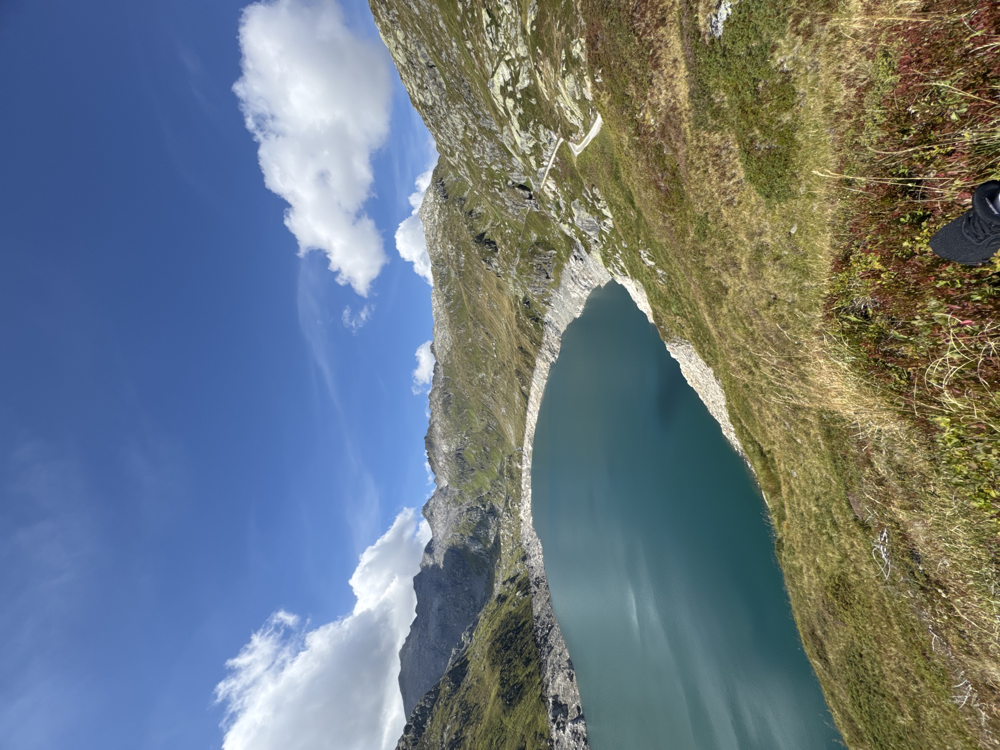 This is a high-angle, wide shot photograph of a man-made reservoir in a mountainous alpine environment. The reservoir's water is a deep turquoise color, contrasting with the rocky, barren shores and the green and brown vegetation covering the surrounding hillsides. The mountains in the background are rugged and rocky, with patches of green grass and some visible snow or light-colored rock on the higher peaks. The sky is a clear blue with scattered white, puffy clouds. A winding dirt path is visible traversing the hillside to the right of the reservoir. The foreground shows a person's feet, wearing black athletic shoes, positioned on a grassy slope dotted with low-lying reddish-brown plants and dry grasses. The overall scene suggests a remote, natural landscape with human intervention in the form of the reservoir. The location is identified as a reservoir near Airolo, Switzerland.