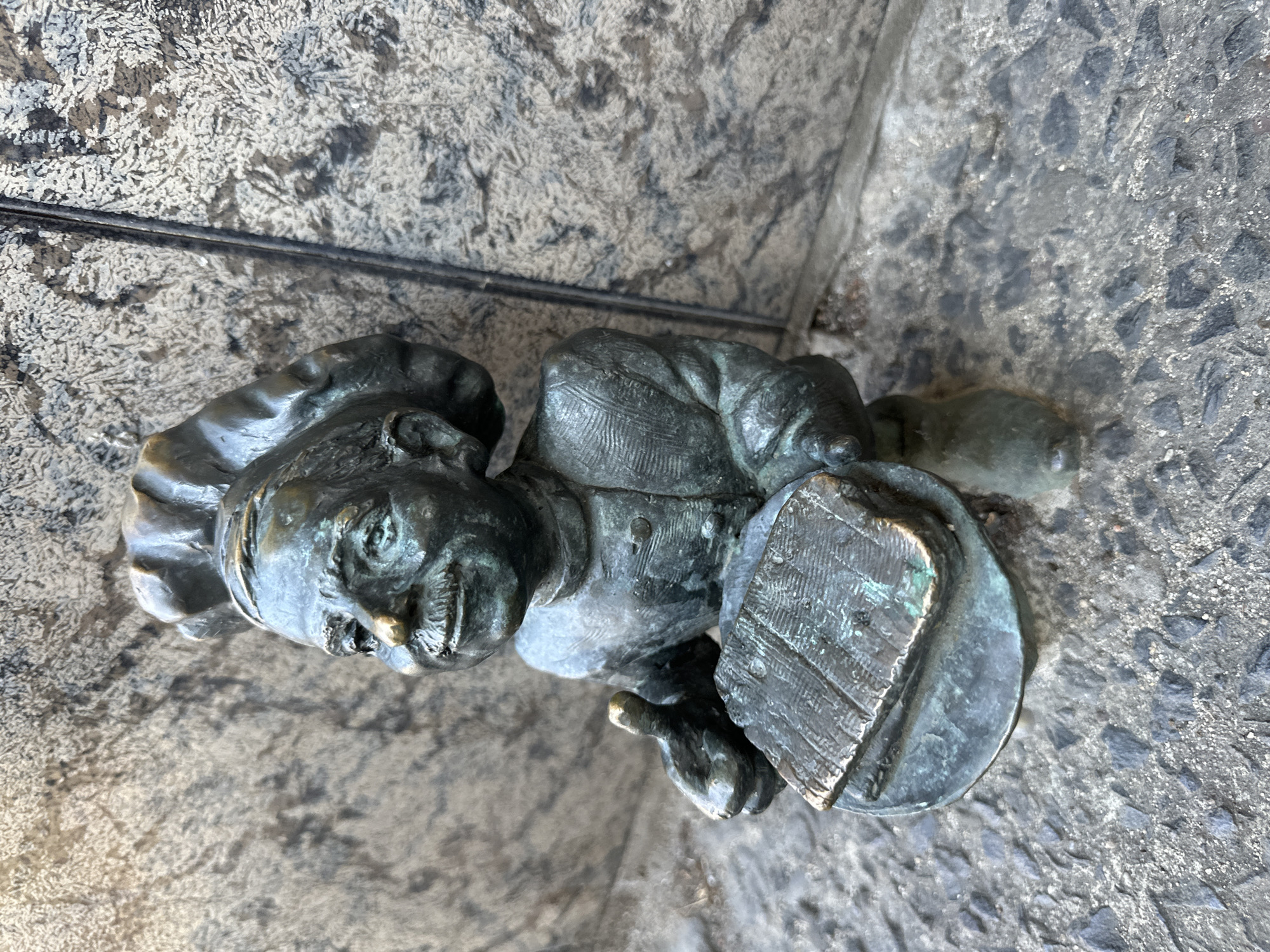 A close-up, slightly angled shot depicts a bronze dwarf statue, a characteristic feature of Wrocław, Poland. The dwarf, wearing a chef's hat, is positioned on a gray, uneven stone surface suggestive of a sidewalk or plaza. The dwarf is holding a large, round object that resembles a frying pan or lid, with what appears to be a cooking utensil laid across it. The metal of the statue has a patinated finish with green and dark gray tones, indicating age and exposure to the elements. A dark, thin object, perhaps a cable or pipe, lies above the dwarf near a light-colored stone wall. The dwarf is looking to the left side of the image. The statue’s overall condition suggests it is a well-worn, possibly touched by passerby, and popular tourist attraction. The specific location within Wrocław cannot be determined from the image alone.