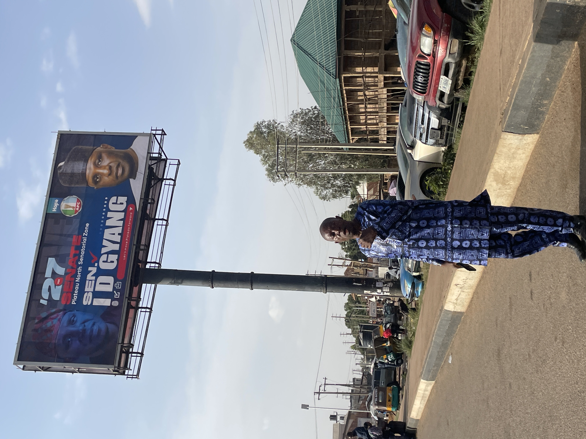 The image shows a man standing on a street in Jos, Nigeria, next to a large billboard advertising Sen. I.D. Gyang for the Plateau North Senatorial Zone. The billboard features two images of Sen. Gyang, one in a traditional cap and the other in a headdress. The text on the billboard includes "27 for Senate" and "Let's move Plateau Forward." The man in the foreground is wearing a blue and white patterned outfit. The street shows parked cars and businesses in the background.