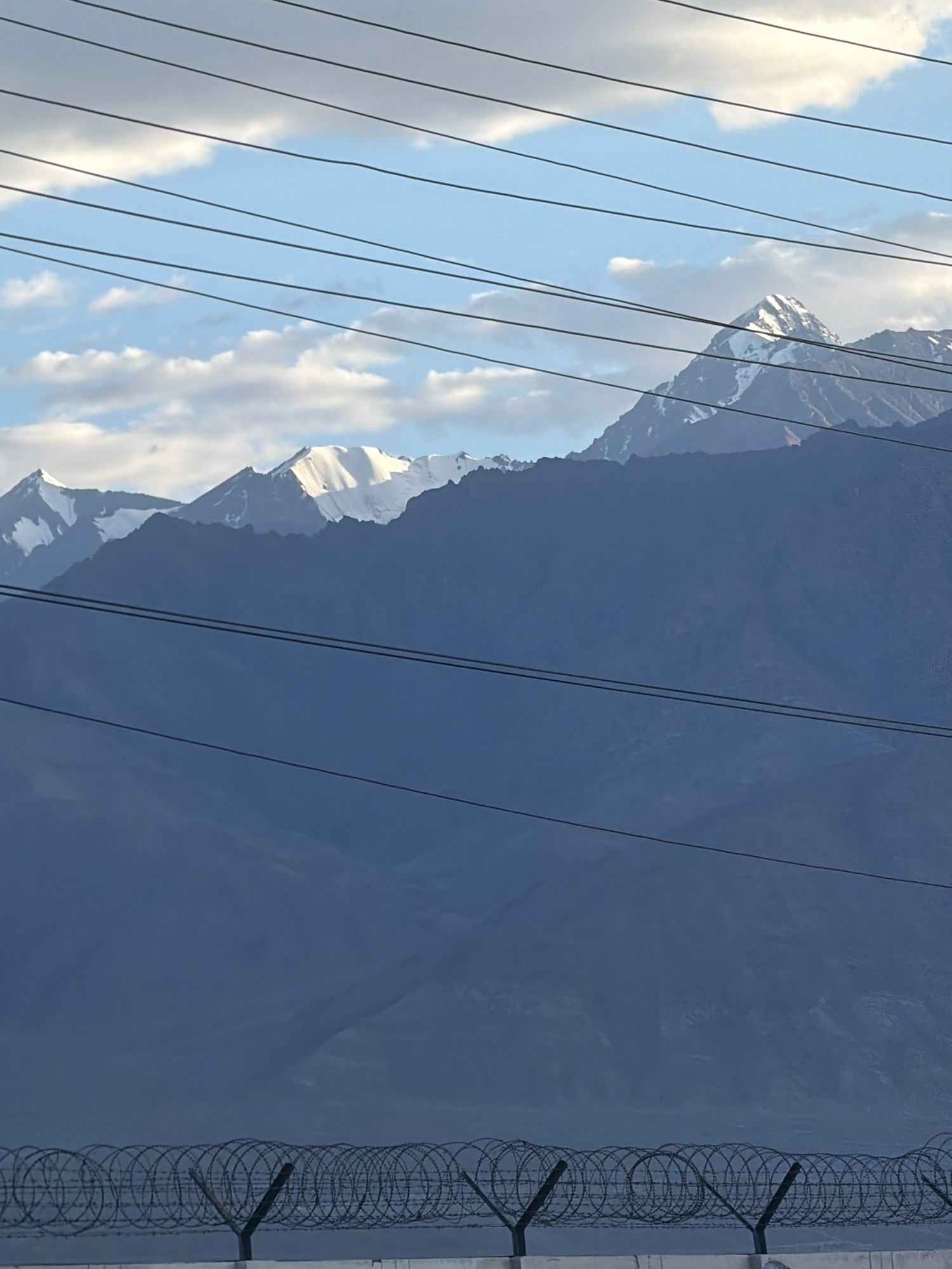 The image depicts a mountainous landscape with snow-capped peaks under a partly cloudy sky. Several parallel power lines stretch across the upper portion of the frame, and a barbed wire fence runs along the bottom. The terrain appears arid and rugged, consistent with the geography of Leh, India, a high-altitude desert region. There are no people or animals visible in the image.