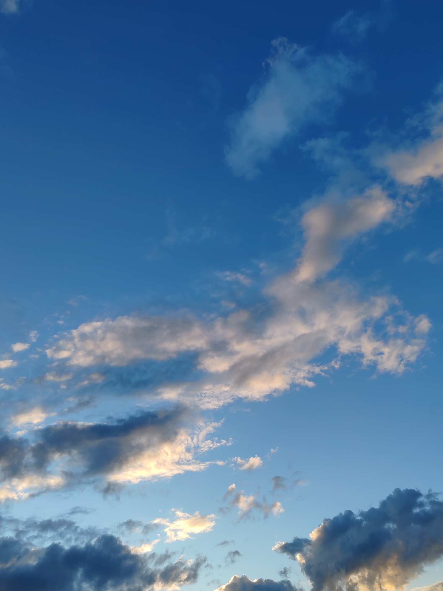 This is a photograph of the sky with clouds, taken during daylight. The sky is blue with varying shades of white, grey, and dark blue clouds scattered throughout. The clouds are illuminated by sunlight, giving some of them a warm, golden hue. The photograph does not contain any people, subjects, objects, actions, or interactions. The location is not discernable from the image itself, and therefore any association with the city of Чухлэм, Russia, cannot be confirmed.