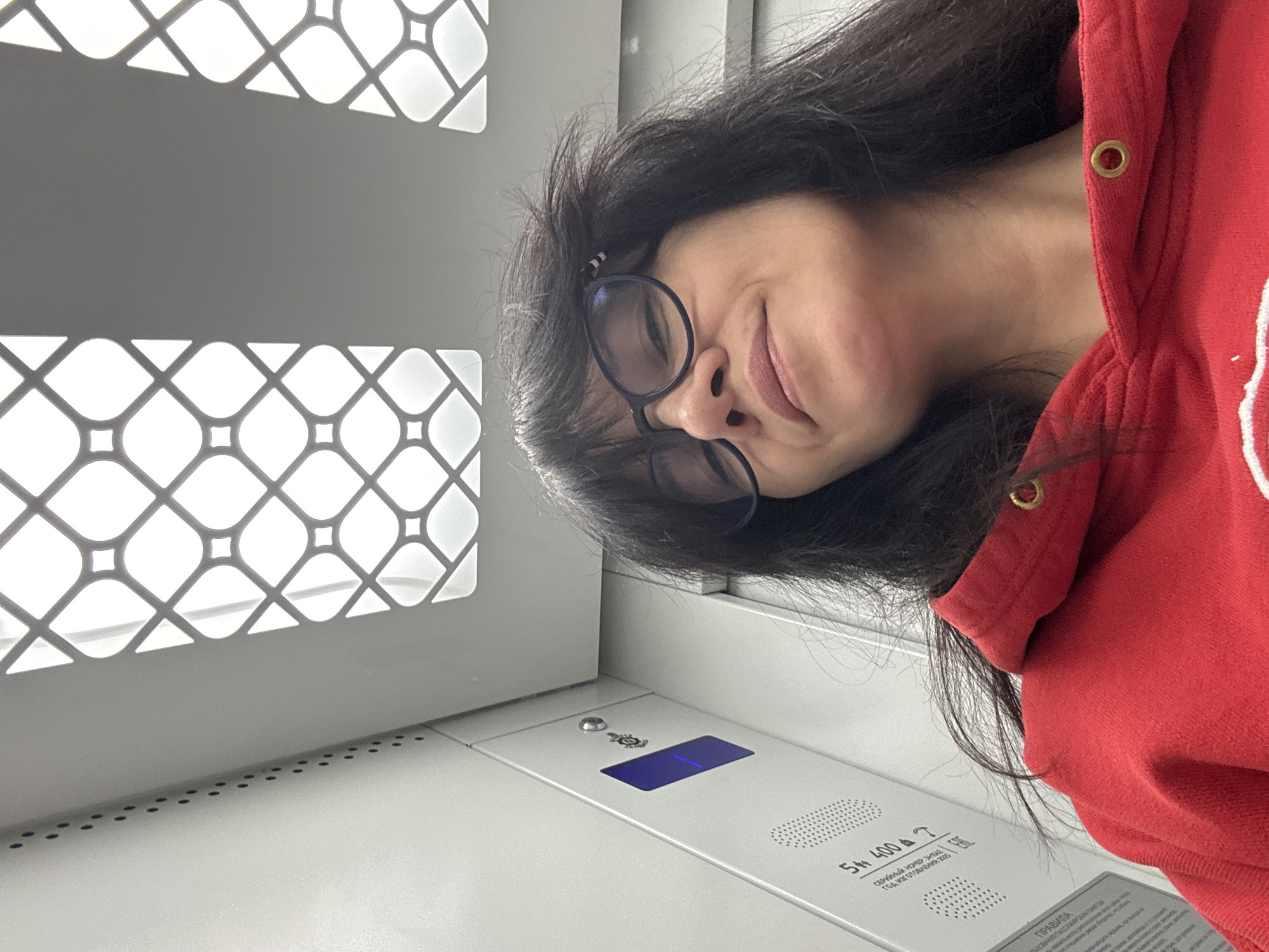 A selfie of a woman wearing glasses and a red hooded sweatshirt in an elevator. The elevator has a patterned metal grate on the wall and a control panel. Text in Russian is visible on the control panel. The city of Chaikovsky, Russia, cannot be confirmed as the location based on the image.