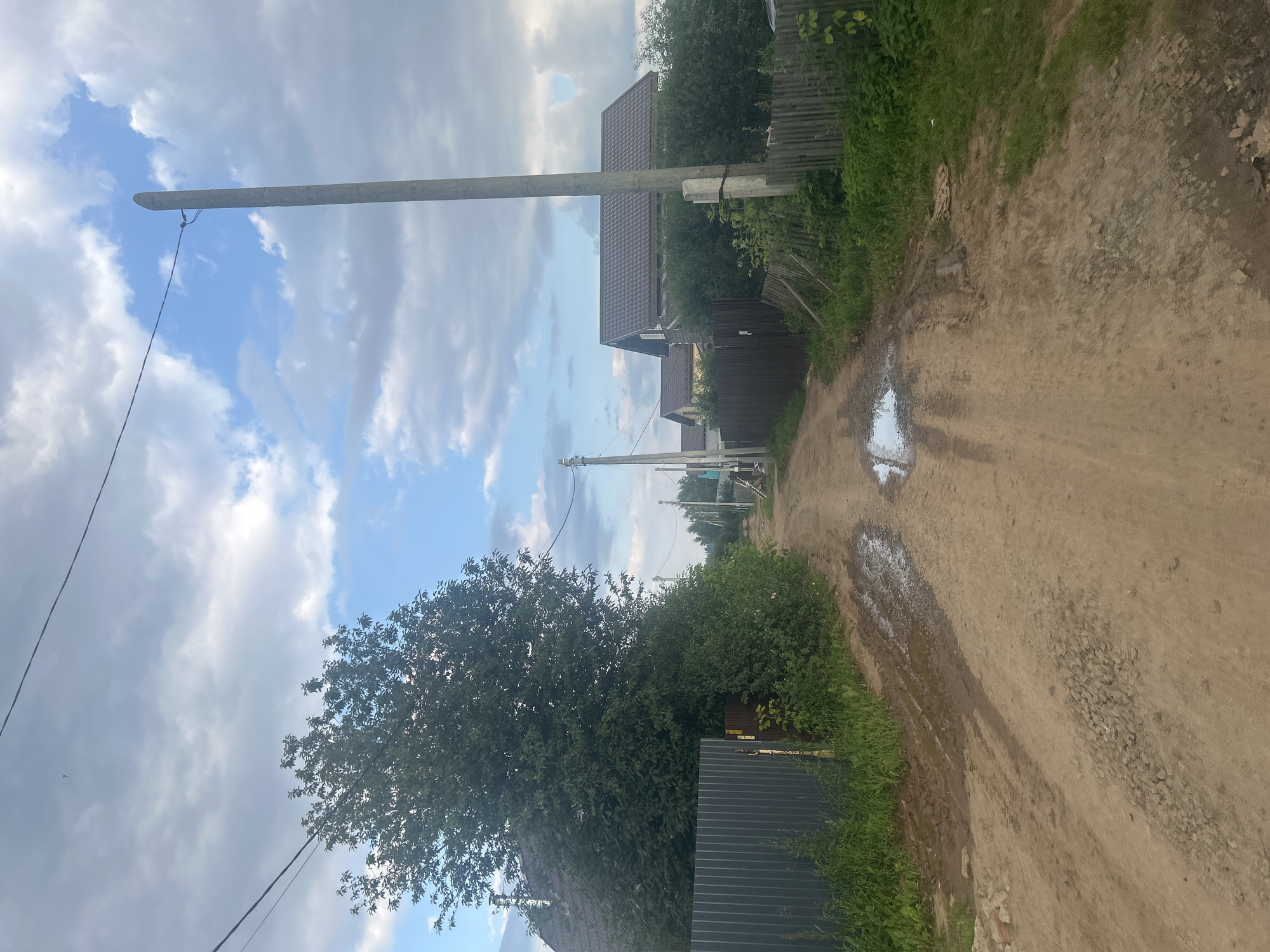 The image depicts a dirt road in a rural setting, possibly the village of Petrovka, Russia. The road is uneven and shows signs of recent rain, with puddles and muddy patches visible. On either side of the road, there are fences and houses with brown roofs. Lush green trees and vegetation line the road, partially obscuring the houses. Power lines and utility poles are present along the road. The sky is partly cloudy, with a mix of blue sky and grey clouds. There are no people or animals visible in the image, nor are there any apparent objects of particular interest beyond the general scene.