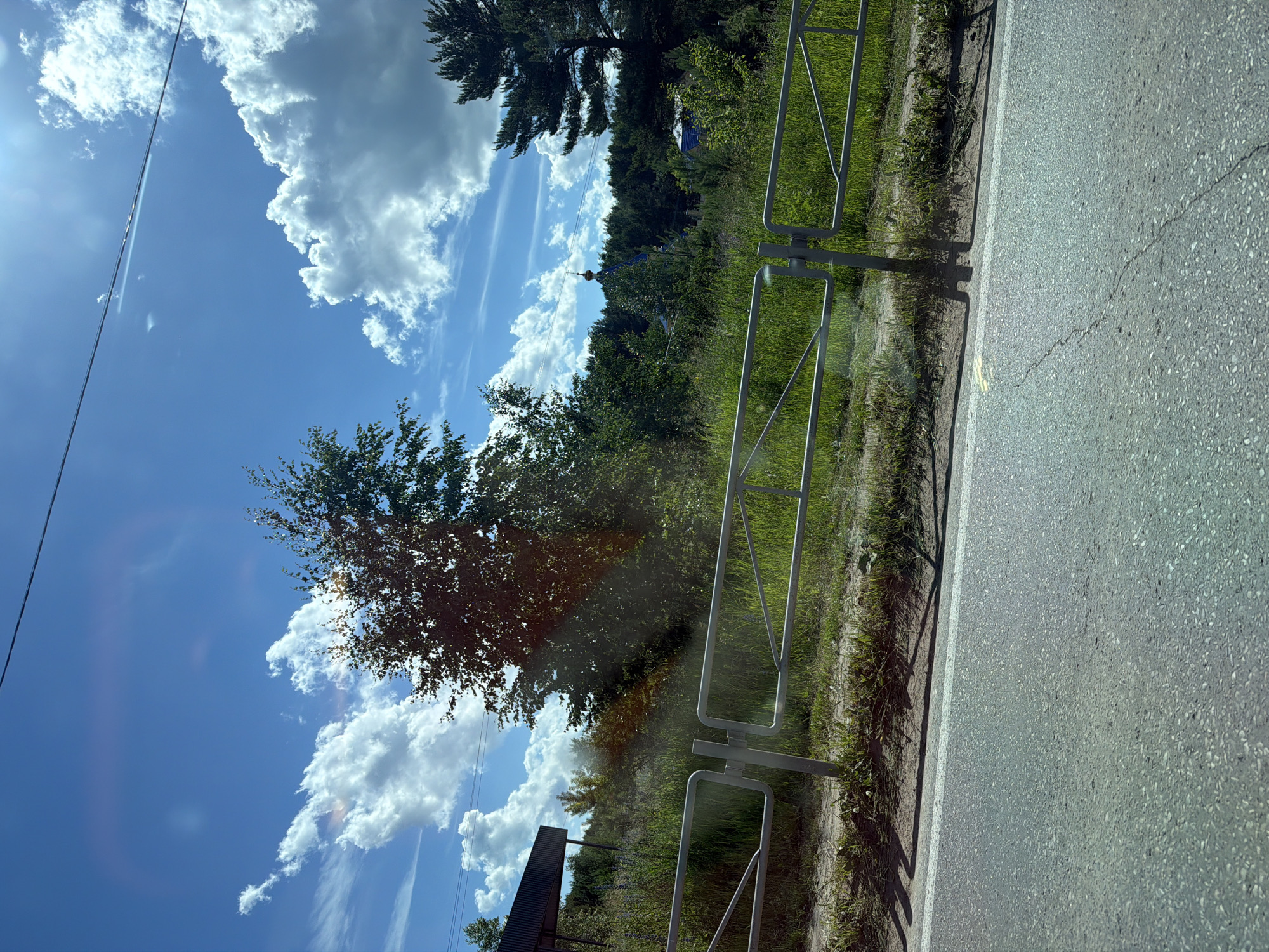 The image shows a view of a rural landscape with a road in the foreground. A metal fence runs along the side of the road, separating it from a grassy area with trees and vegetation. In the background, a glimpse of a blue-domed building with a cross, likely a church, is visible amidst the trees. The sky is blue with scattered white clouds, and the sun is shining, casting shadows. A power line runs across the top of the image. The scene appears to be in a rural area, consistent with the outskirts of Chaikovsky, Russia, characterized by its natural scenery and presence of religious architecture. No people or specific actions are depicted.