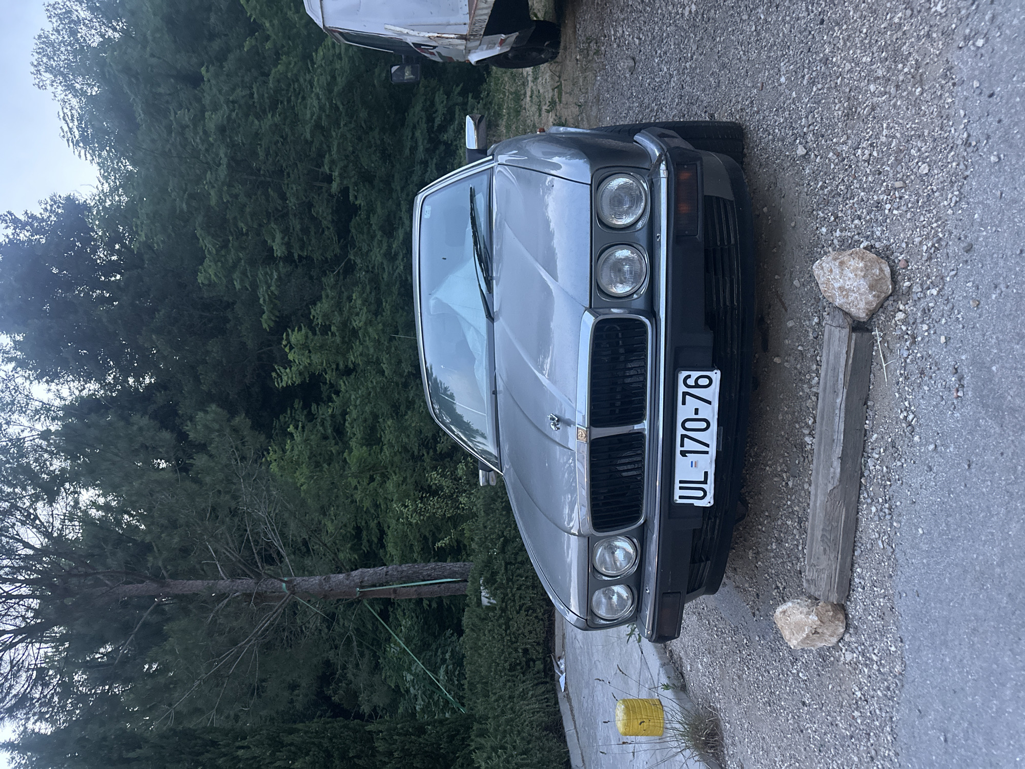 The media features a classic silver or light grey Jaguar XJ (likely an XJ40 series) luxury sedan parked outdoors. The car is equipped with distinctive stacked circular headlights and a prominent grille featuring the Jaguar "leaper" mascot on the hood.
The setting appears to be a semi-rural or natural environment, with a ground surface composed of both paved asphalt and gravel. Lush, dense green trees and foliage form a significant backdrop behind the vehicle. In the mid-ground, a tree with a green strap around its trunk is visible.
No specific activity is taking place; the car is stationary. The time of day appears to be daytime, possibly early morning or late afternoon, given the soft, diffused light. The weather seems clear, with a bright sky partially visible above the trees.
Notable details around the car include a large, weathered wooden beam lying on the ground parallel to the front bumper, secured by a natural rock. Another rock is present nearby. A yellow object, resembling a container or bucket, is partially visible on the ground to the lower left. In the background, another older, white vehicle (possibly a truck or van) is partially visible behind the Jaguar.
The most prominent visible text is on the car's license plate, which reads "UL 170-76." The "UL" prefix indicates that the car is registered in the Ulcinj region of Montenegro, aligning with the provided location context of Boreti, Montenegro.